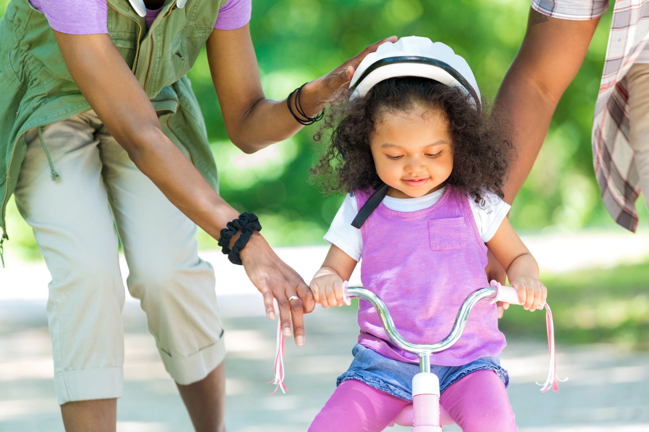 Beautiful African American daughter being taught how to ride a tricycle. Her parents arms are in the shot as they help keep her on the bike. She is wearing light pinks and purples. She has beautiful black curly hair.