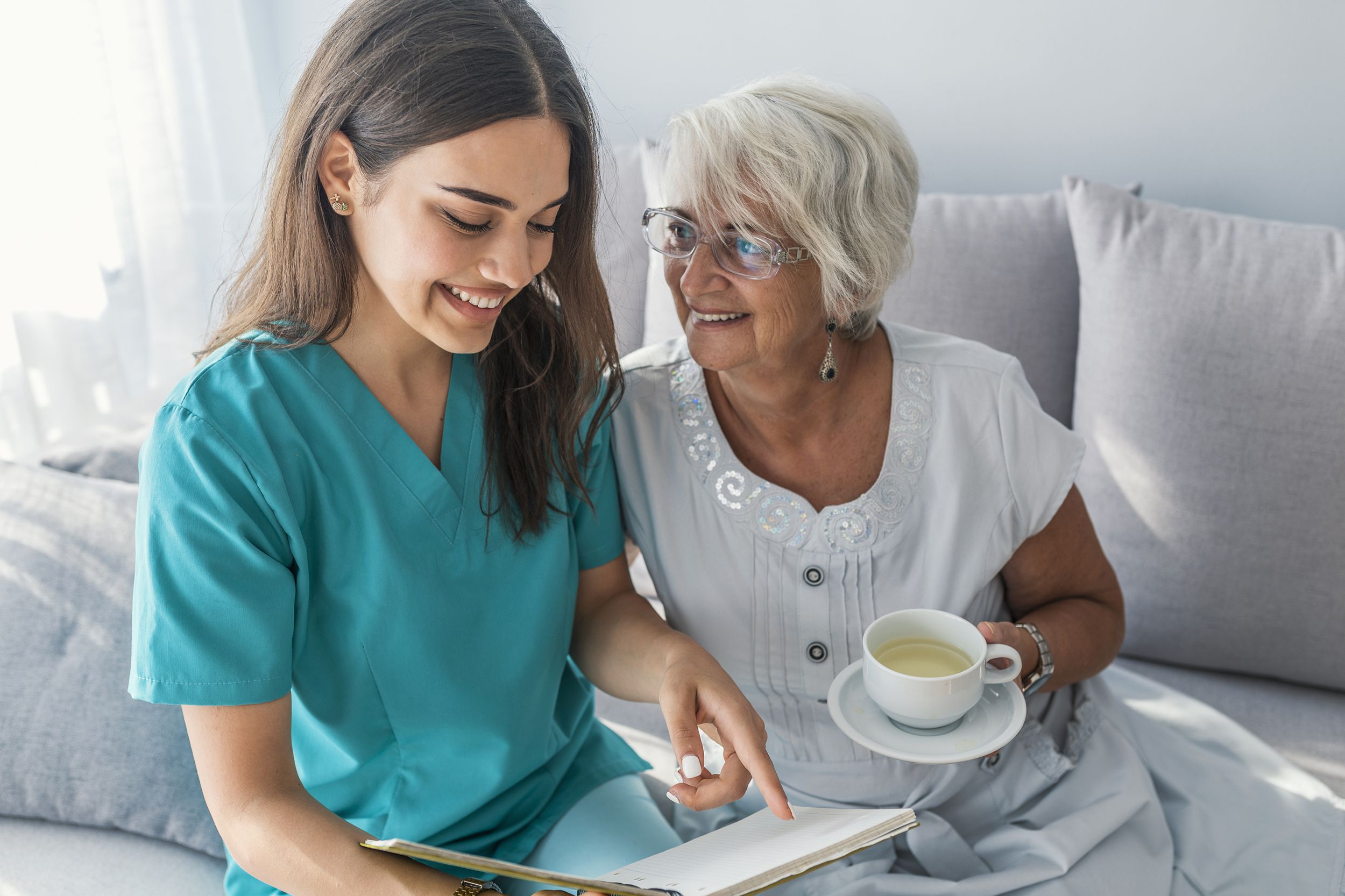 happy elder woman sitting on white sofa and listening to nurse reading a book out loud