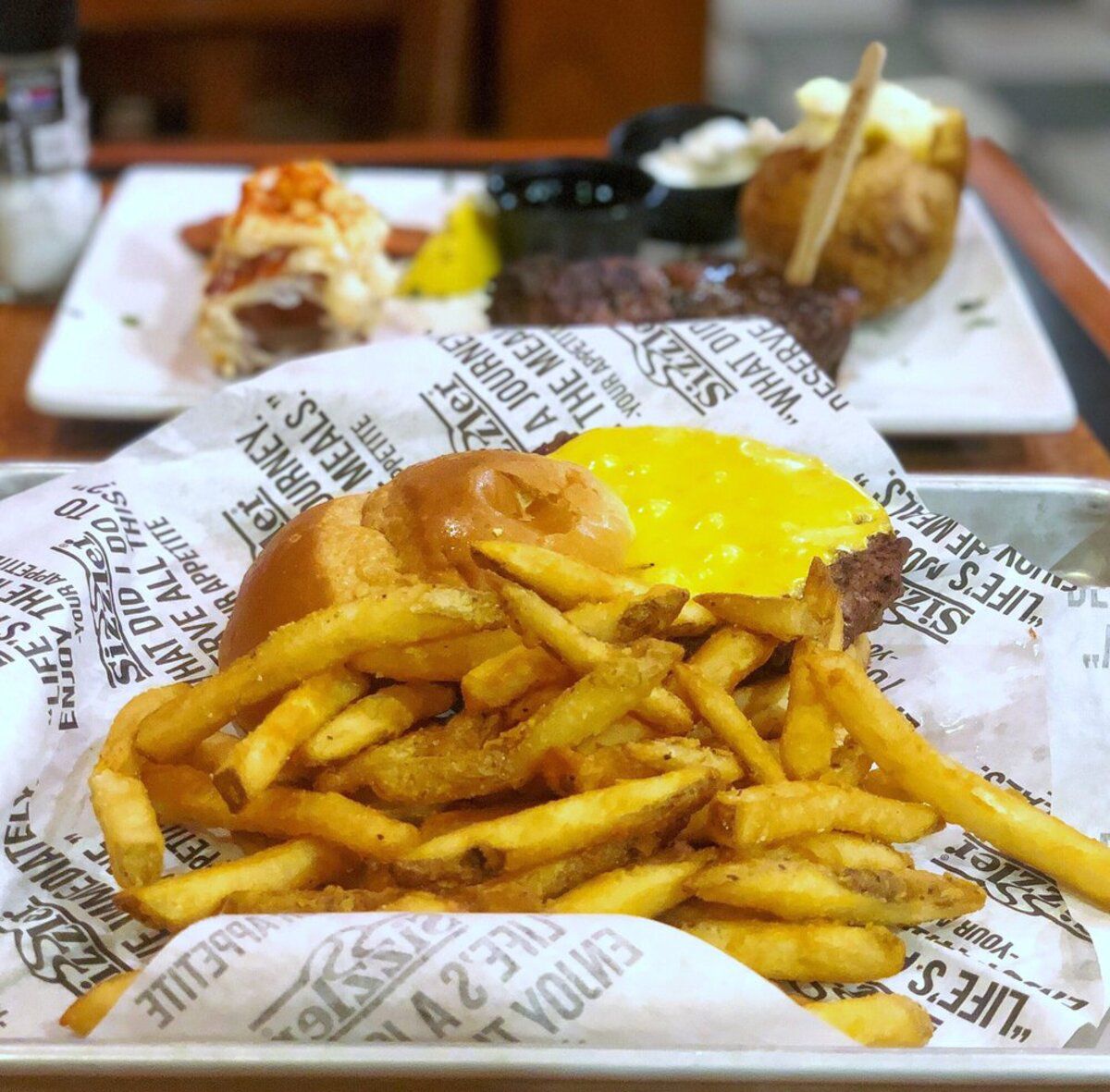 hamburger with fries on plate in foreground and steak with sides on plate in the background on table at Sizzler, Santa Clara, California