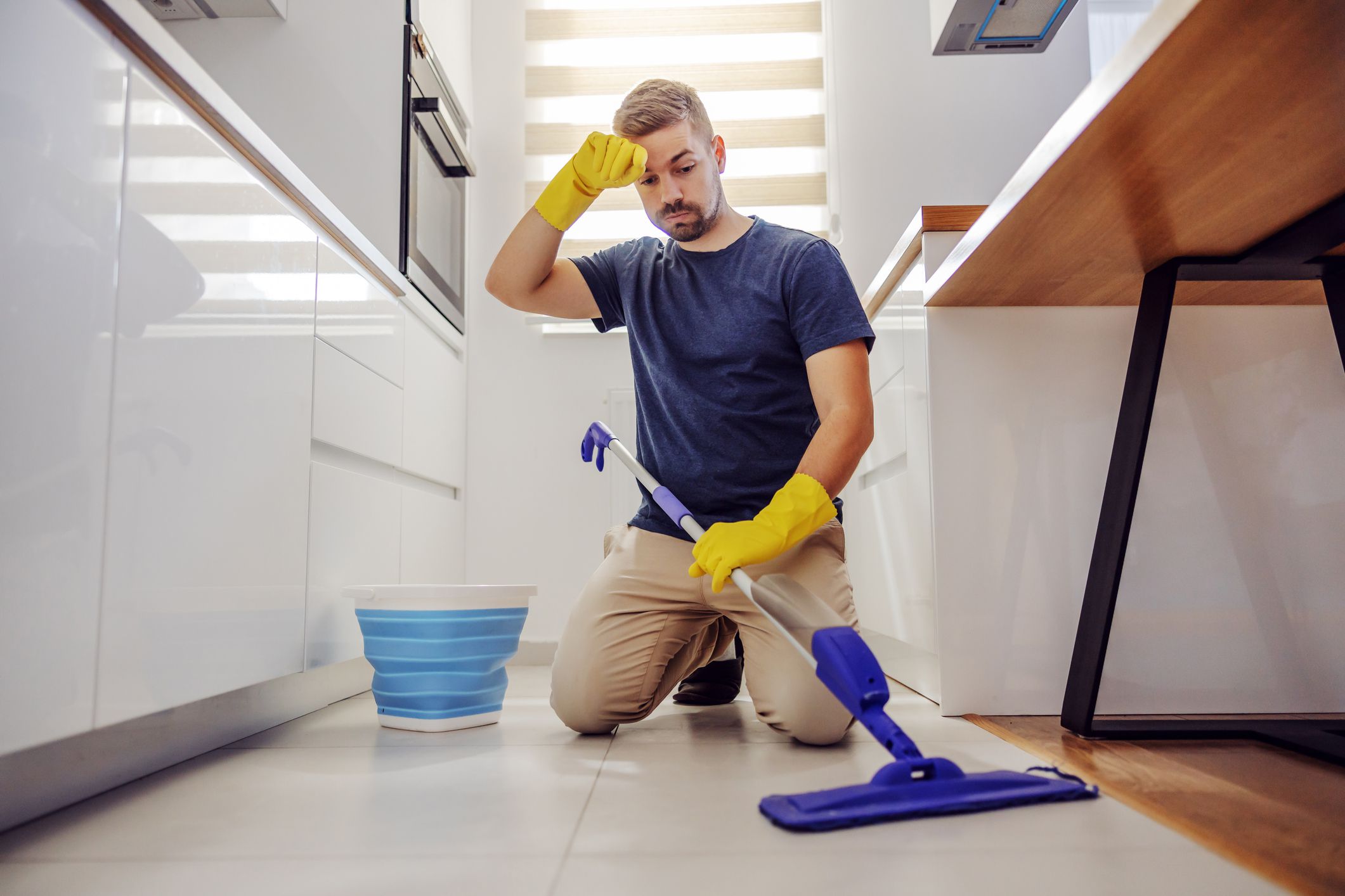 Young tidy worthy tired bearded man kneeling, wiping sweat and trying to clean dirty floor in kitchen.