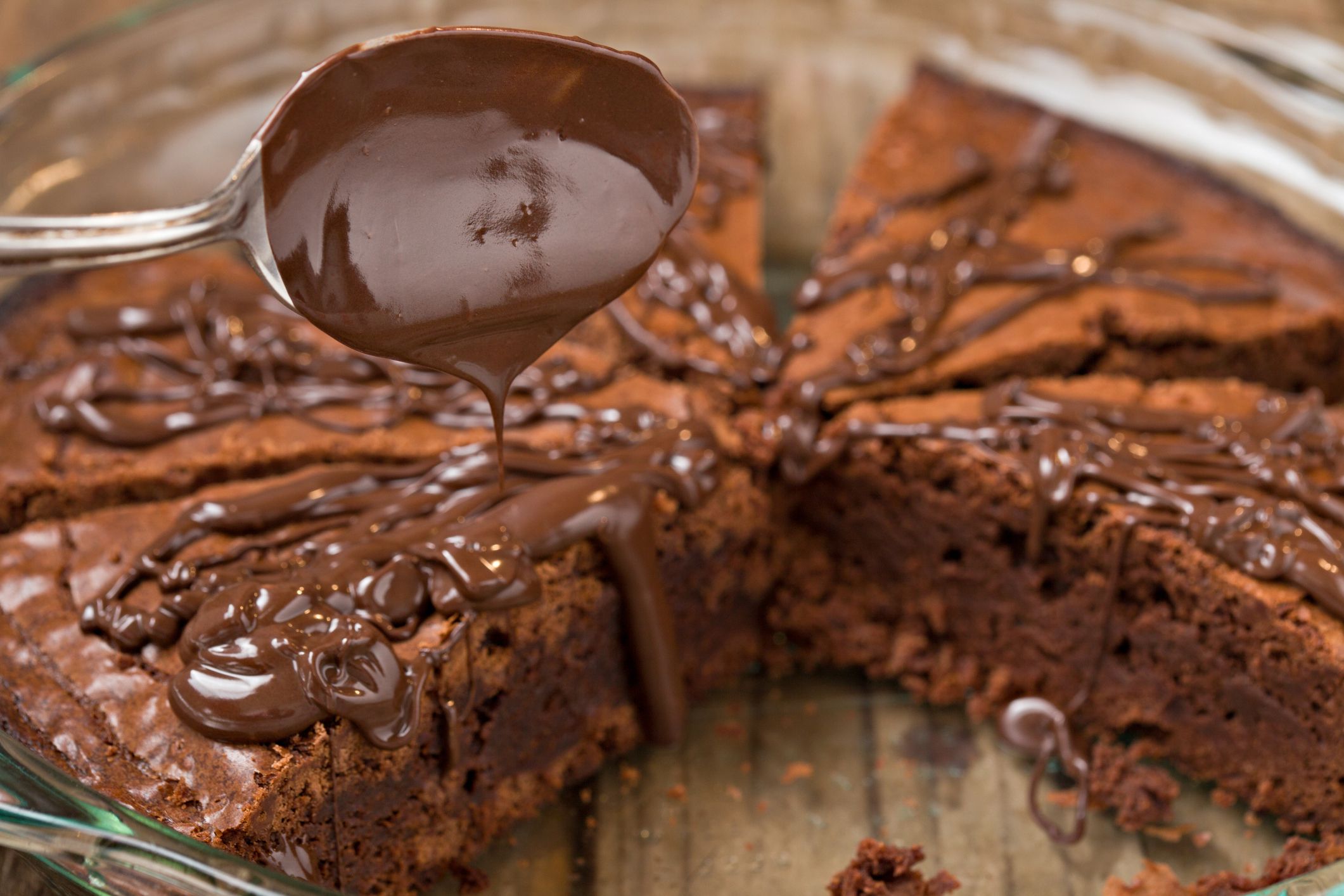 A close up shot of chocolate fudge ganache being poured on freshly baked brownies in a round glass baking tray.