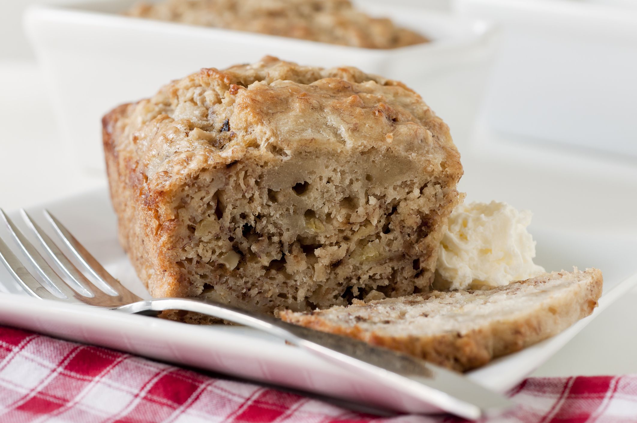 Mini sliced loaf of banana bread with a dollop of whipped cream and fork on a square white plate. A second mini loaf in a loaf pan is visible in the background.Similar Images: