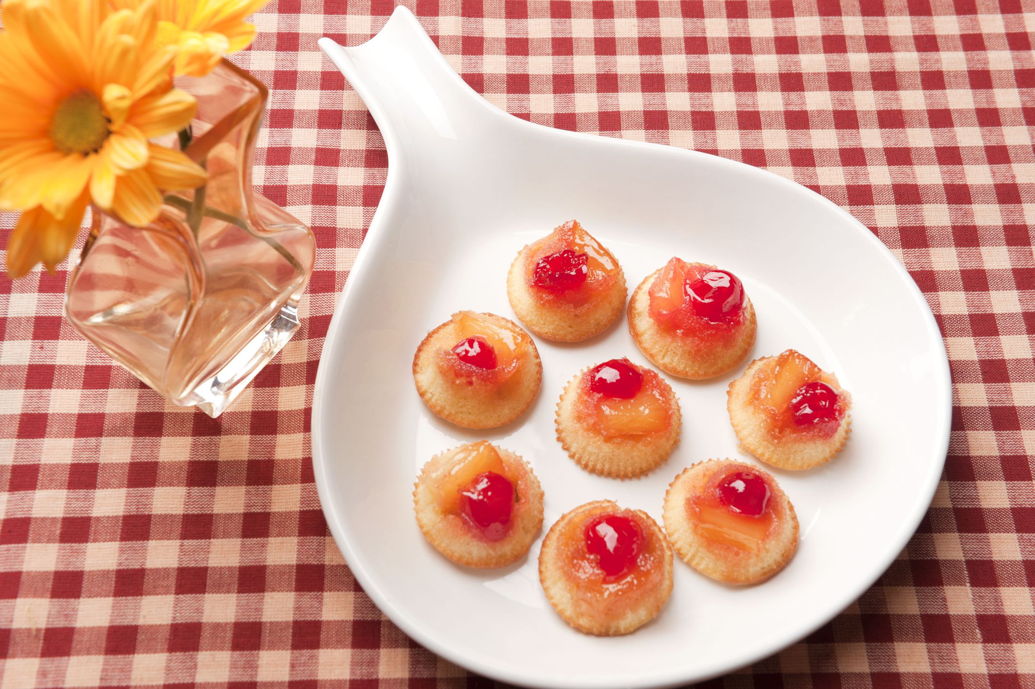 Bite sized pineapple upside down cake on a white pan next to an antique vase of golden yellow daisies. Studio Shot.