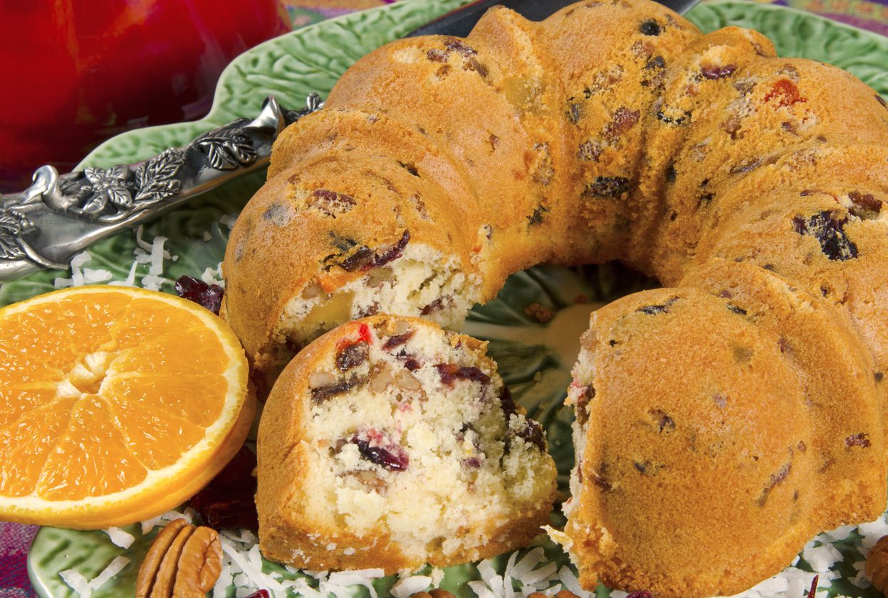 This is a Fruit and Nut Cake with an orange slice and pecan for decoration. One slice has been cut out and is displayed laying on its side. It is sitting on a formal, green and patterned place setting with a red background. It includes the lower portion o