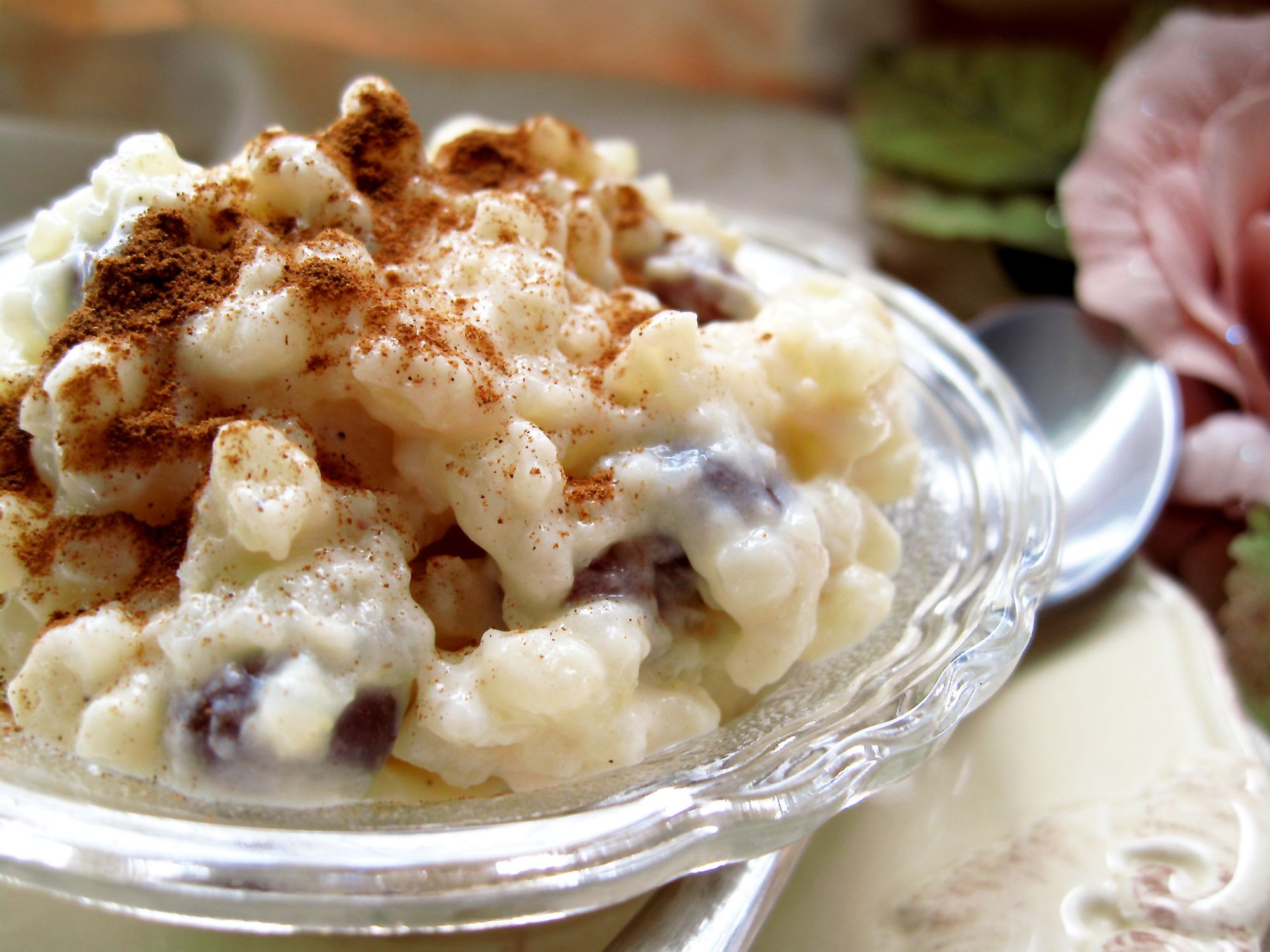 A rich and creamy rice pudding in a glass dish on plate. Cinnamon and raisins, pink rose, spoon.