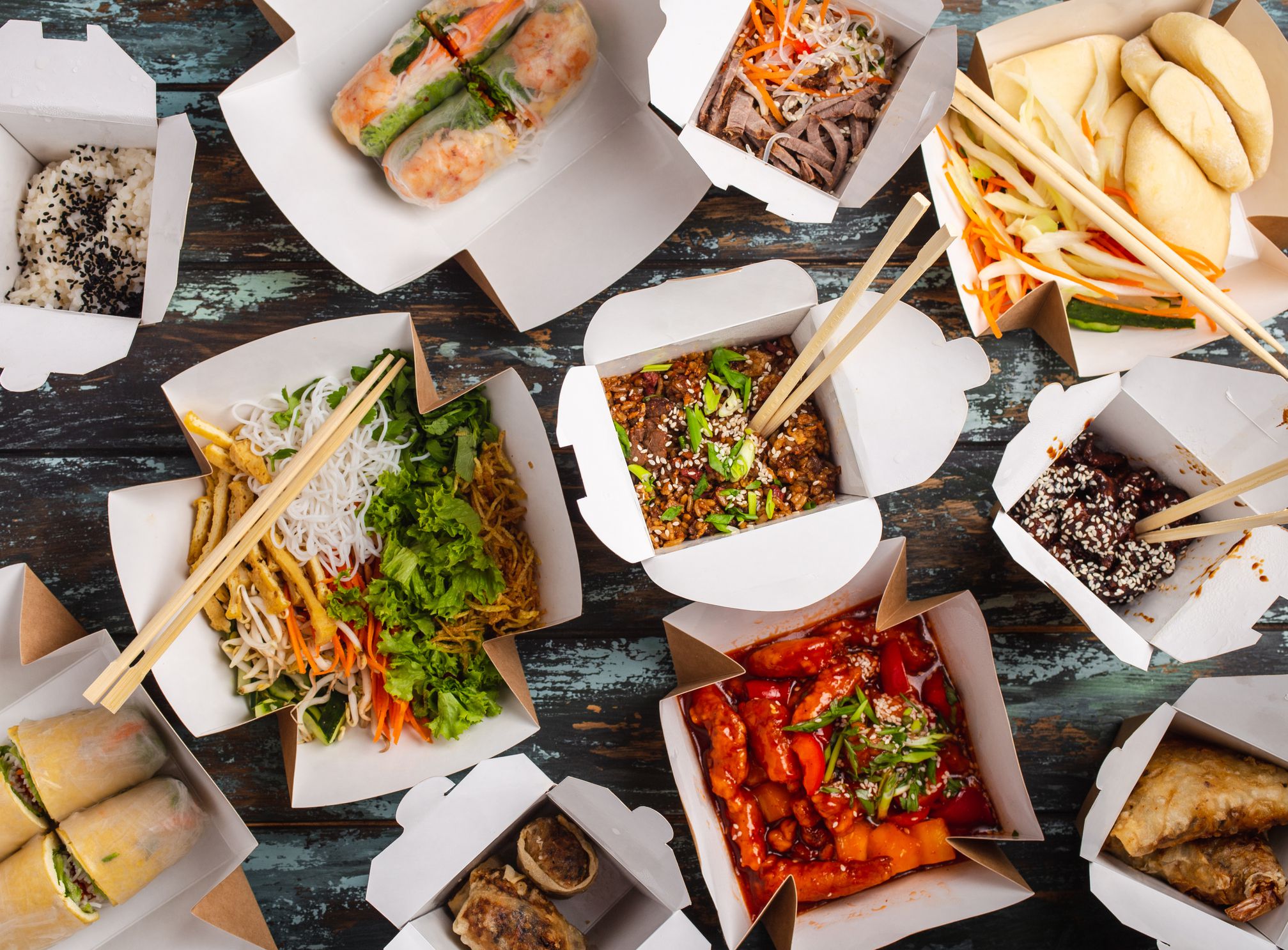 Assorted Chinese dishes in paper delivery boxes: sweet and sour chicken, dim sum, spring rolls, noodles, salad, rice, steamed buns, dips. Asian restaurant take away concept, top view