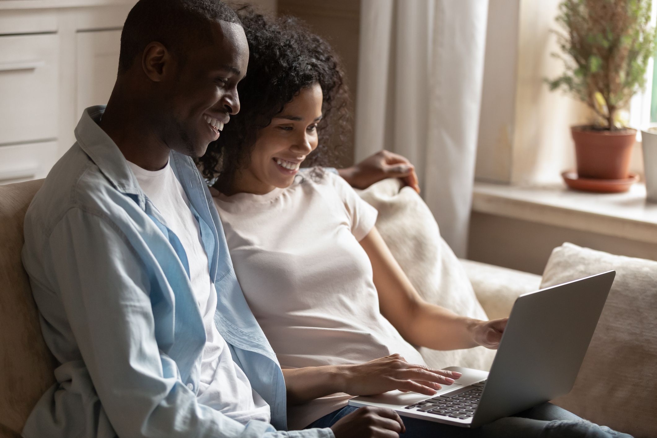 Positive african 30s couple sitting on couch using pc looking at laptop screen enjoy weekend mixed race girl black guy watching online programs videos downloaded movie, lazy activities at home concept