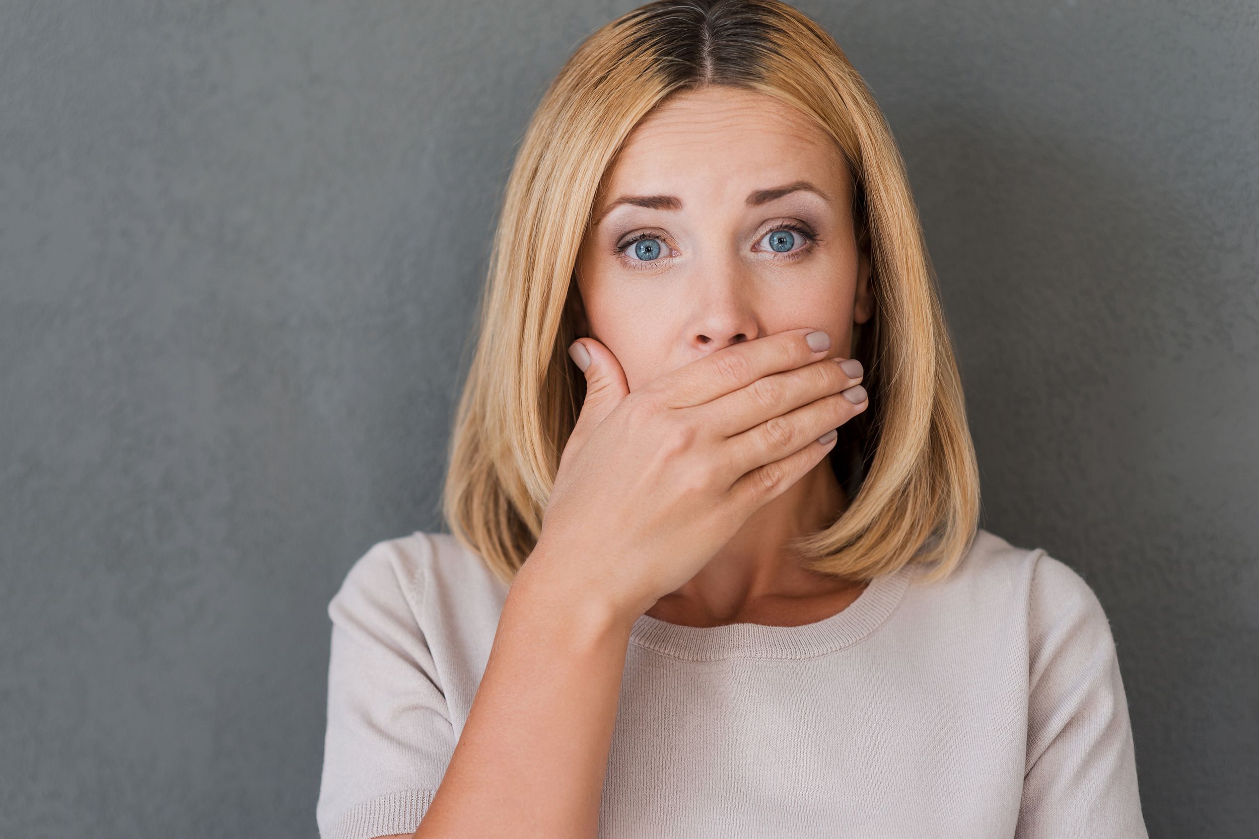 surprised mature woman covering mouth with hand and staring at camera while standing against grey background