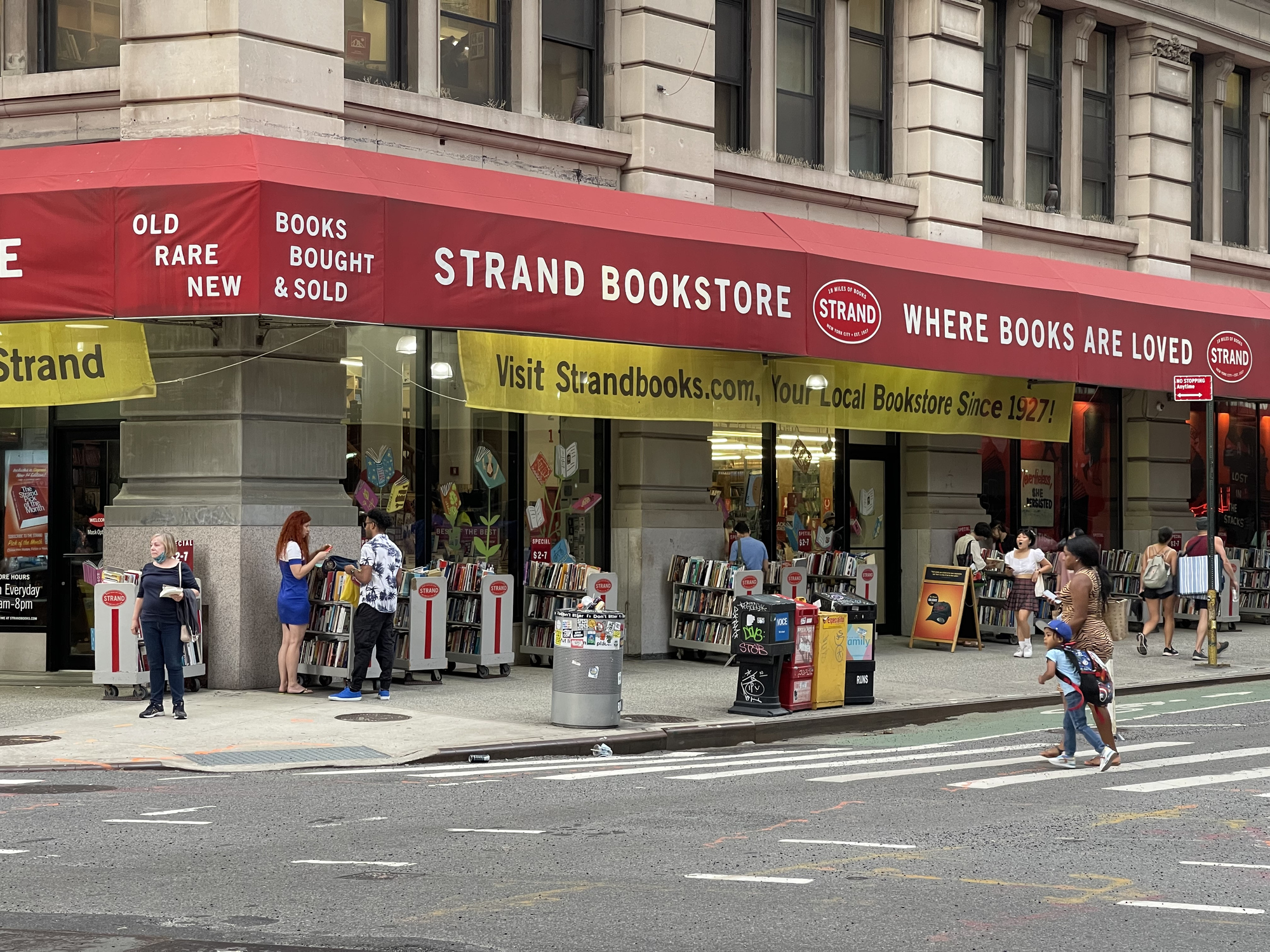 Front exterior of the Strand Book Store, New York City, a few people going through racks of books on the sidewalk, at the corner of 13th Street and Broadway in Manhattan, the road in the foreground