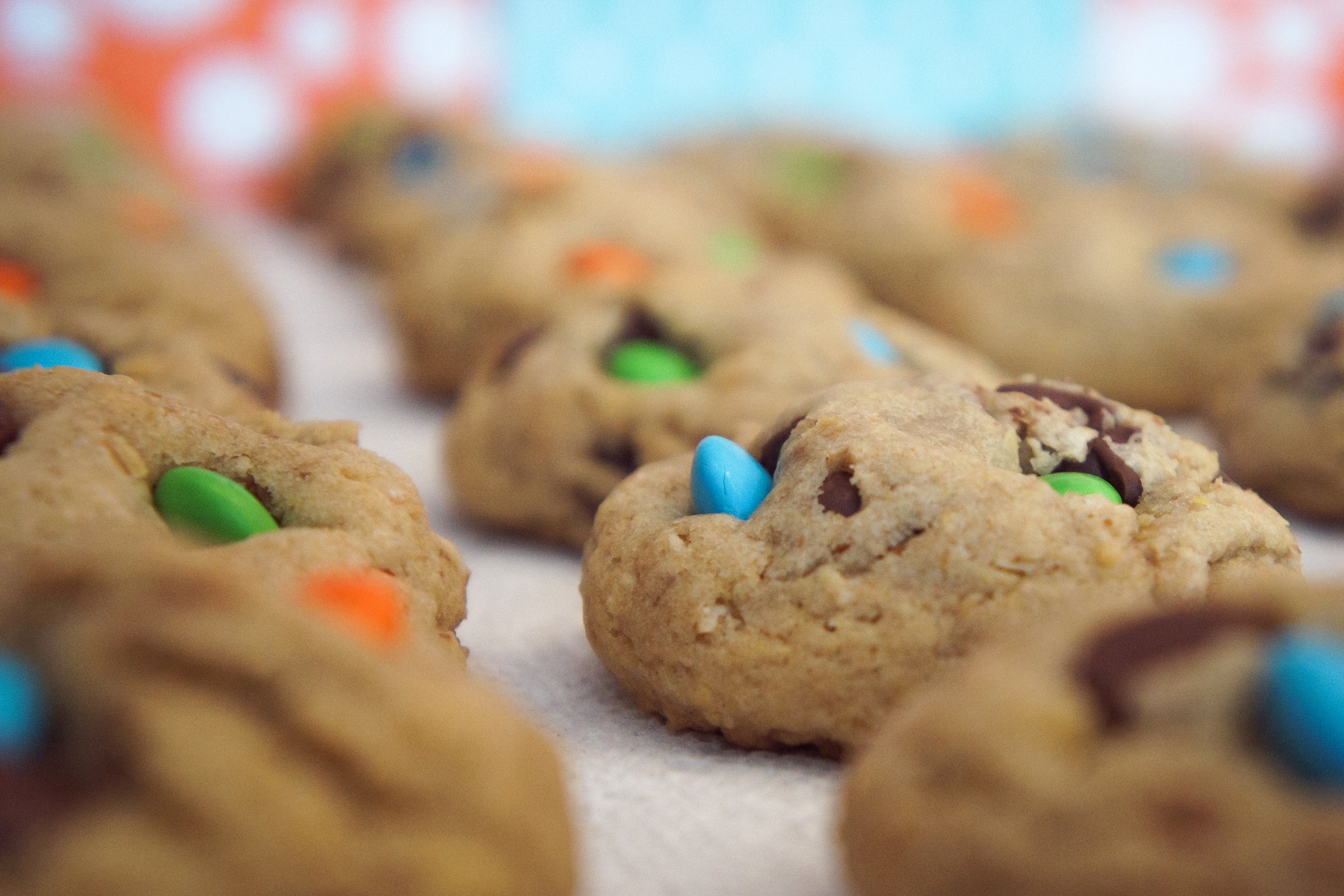 Close-up of cookies with colorful candy-coated chocolates, such as M&Ms, embedded in them. The cookies are placed on a white surface with a blurred, bright background.
