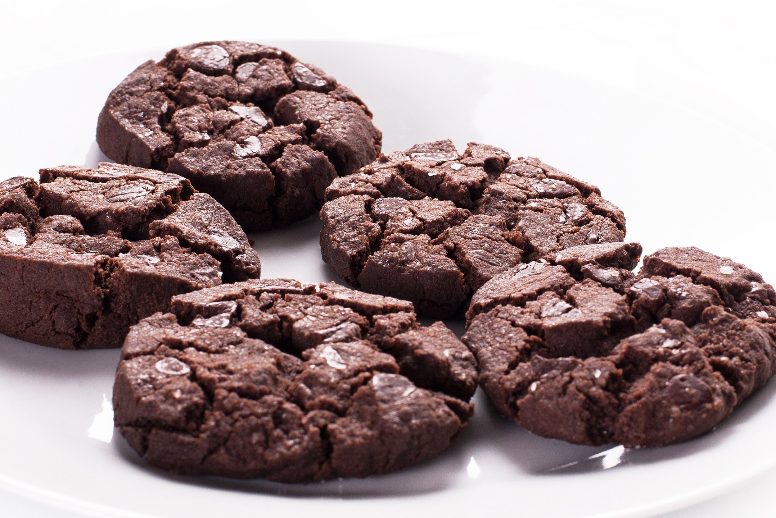 Five chocolate cookies with visible chocolate chips are arranged on a white plate. The cookies have a cracked surface and a rich, dark color, suggesting they are double chocolate or chocolate chunk cookies.