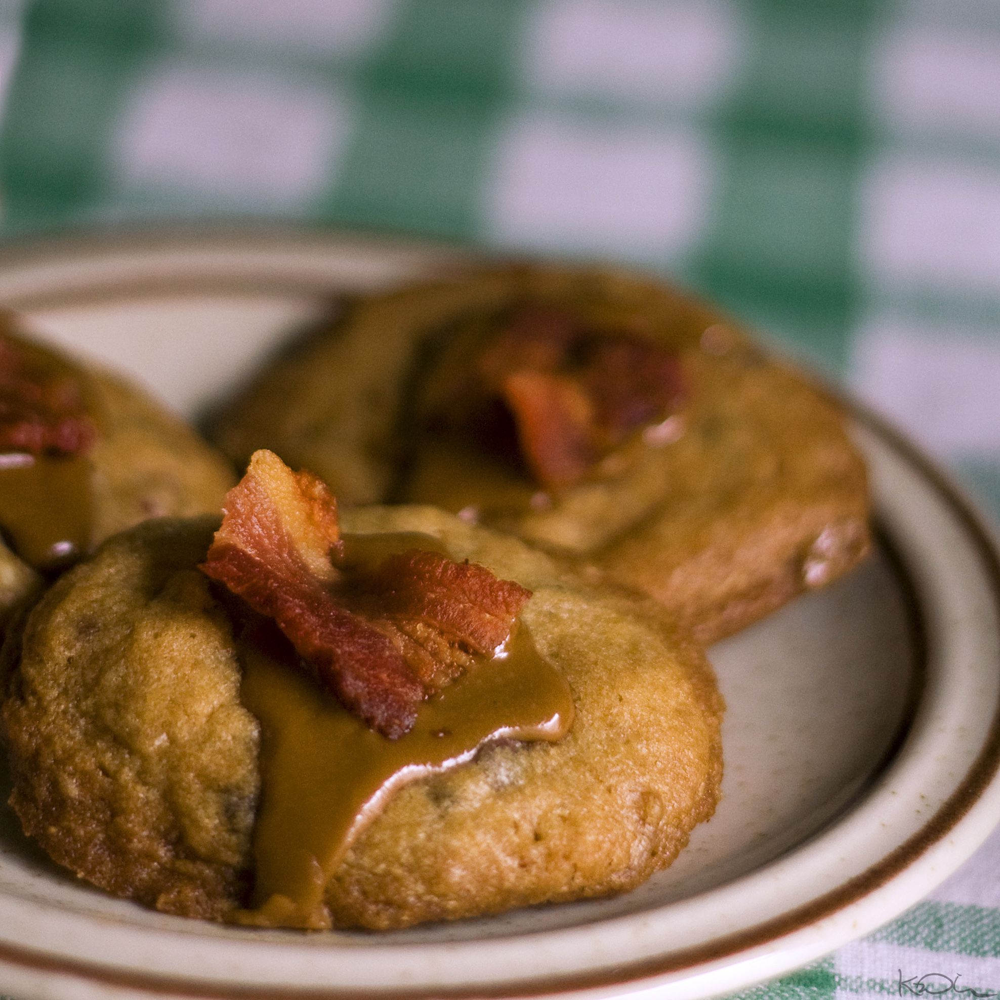 Three cookies topped with caramel sauce and pieces of crispy bacon are arranged on a plate, set against a green and white checkered tablecloth background.