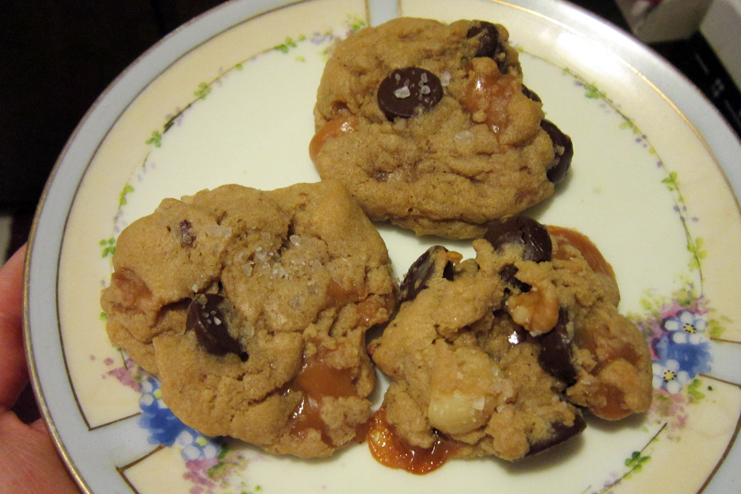 Three homemade chocolate chip cookies with visible caramel chunks and sea salt are arranged on a decorative plate with a floral pattern.