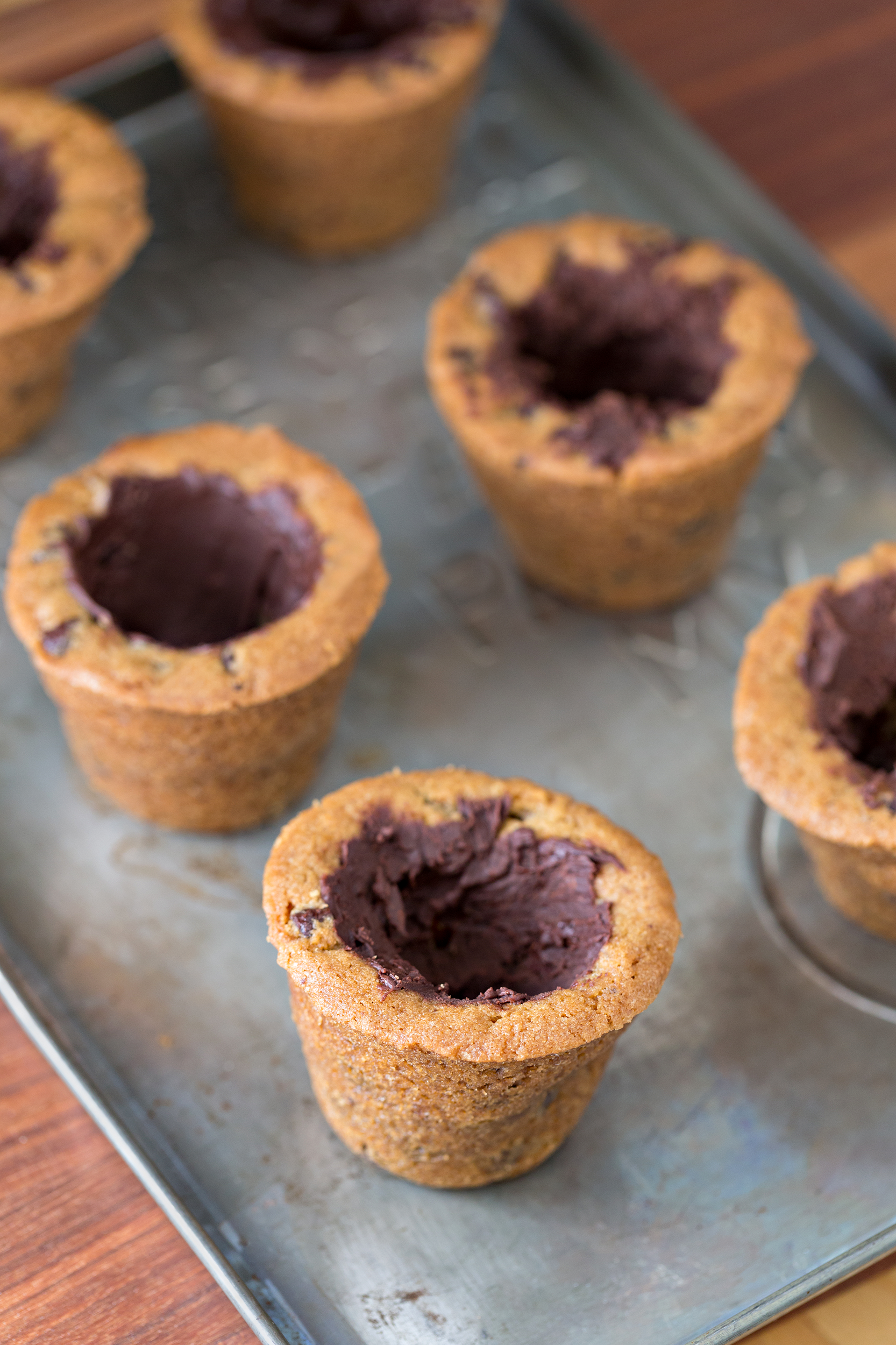 Chocolate chip cookie cups filled with chocolate ganache are arranged on a metal baking tray, with a wooden surface visible in the background.