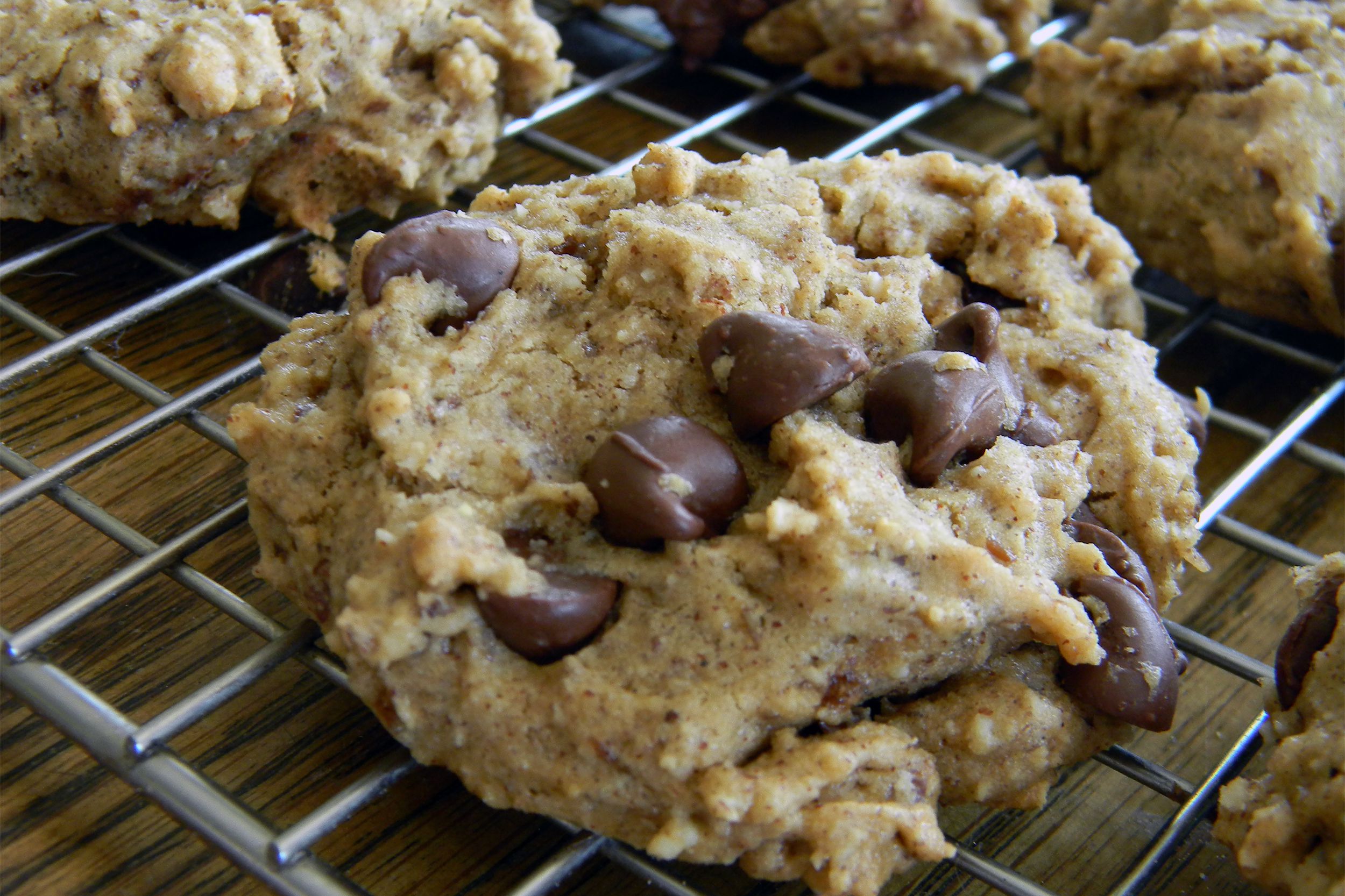 A close-up of a chocolate chip cookie with a soft, chunky texture, cooling on a metal wire rack. Several chocolate chips are visible on the surface of the cookie.