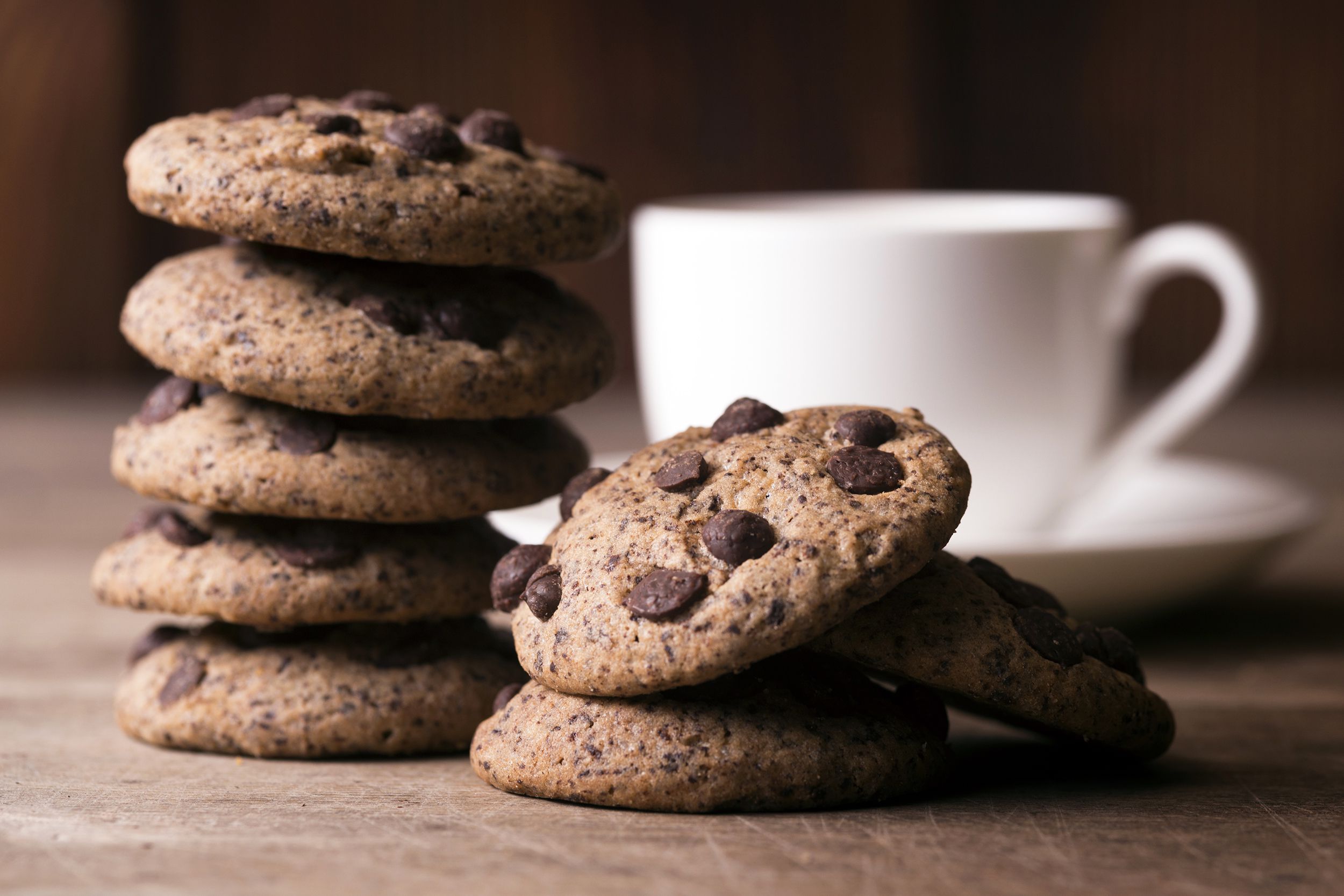 A stack of chocolate chip cookies is arranged on a wooden surface next to a white cup and saucer, with two cookies lying in front of the stack. The background is softly blurred.