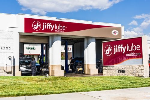 A Jiffy Lube Multicare auto service center with large red signs, open garage bays, workers servicing cars, and a clear sky above. One car is visible inside the garage area.