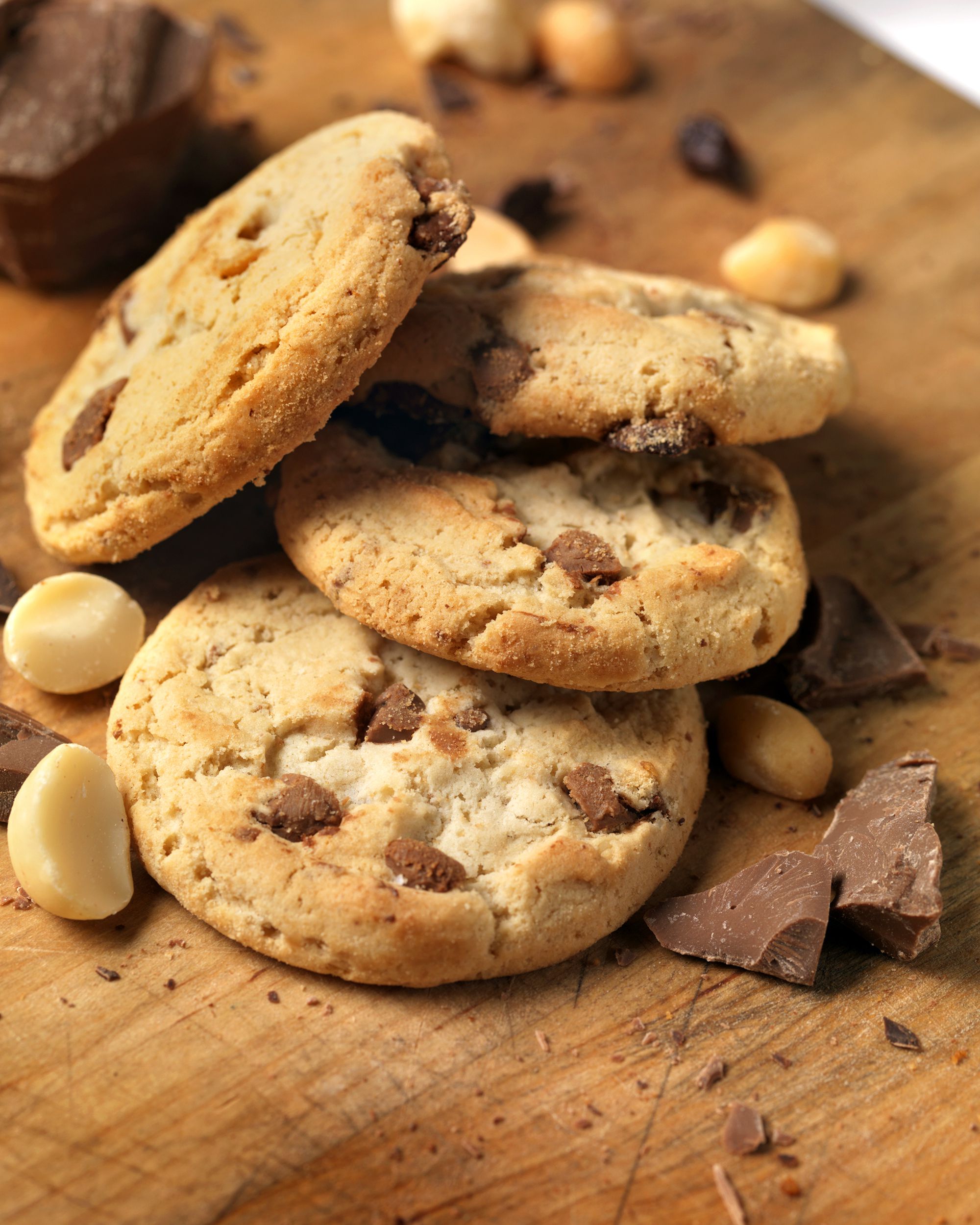 Three chocolate chip cookies stacked on a wooden board, surrounded by pieces of chocolate and macadamia nuts.
