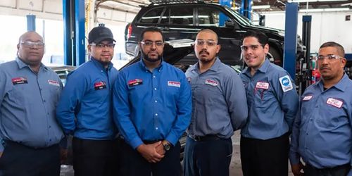 Six auto mechanics wearing blue uniforms and safety glasses stand in a row inside a garage, with a black SUV elevated on a lift and various workshop equipment visible in the background.