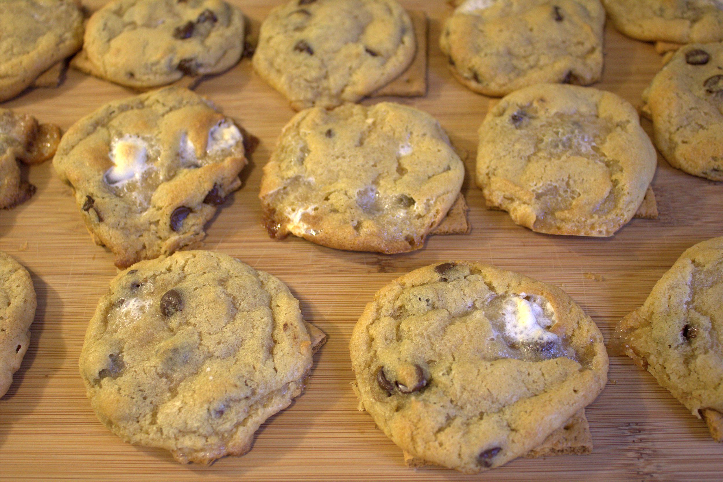 Chocolate chip cookies with visible bits of marshmallow are laid out on a wooden surface, with some cookies placed on square graham crackers.