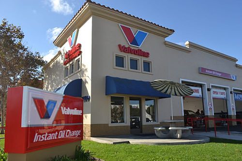 A Valvoline Instant Oil Change service center with a prominent red and white sign in front, blue awnings over windows, and open garage bays on the right side. There is also a round umbrella with a picnic table outside.
