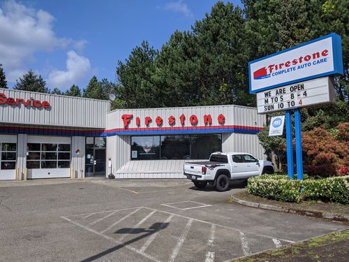 A Firestone Complete Auto Care center with a white pickup truck parked in front. The sign indicates business hours and trees are visible in the background under a partly cloudy sky.