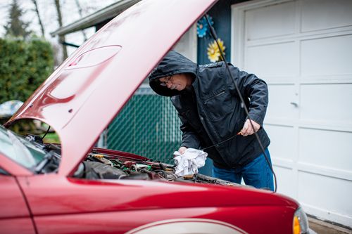 A person wearing a black hooded jacket checks the oil level of a red car with the hood open, wiping the dipstick with a cloth in a driveway near a garage.