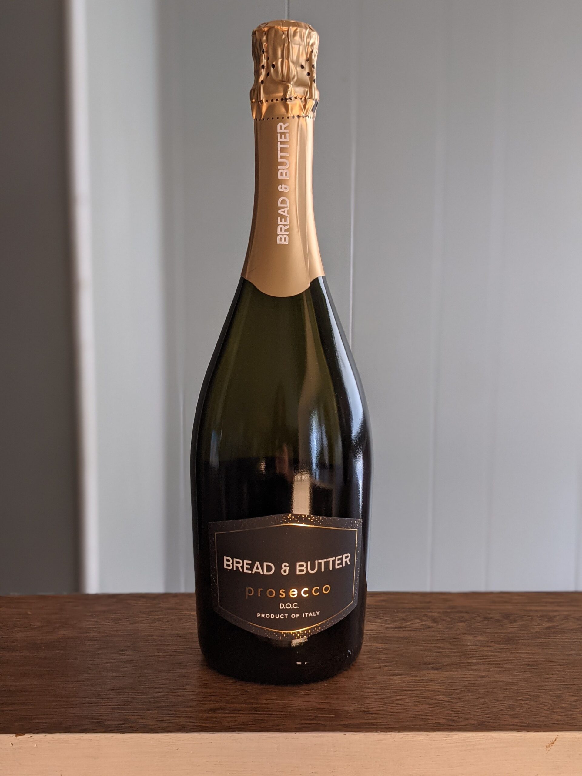 A sealed bottle of Bread & Butter Prosecco on a wooden ledge, white panel wooden wall blurred in the background