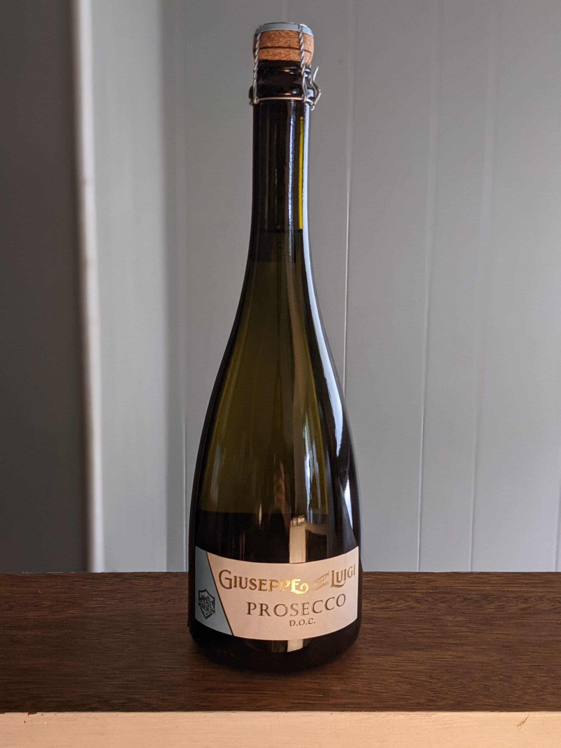 A sealed bottle of Giuseppe & Luigi Prosecco on a wooden ledge, white panel wooden wall blurred in the background