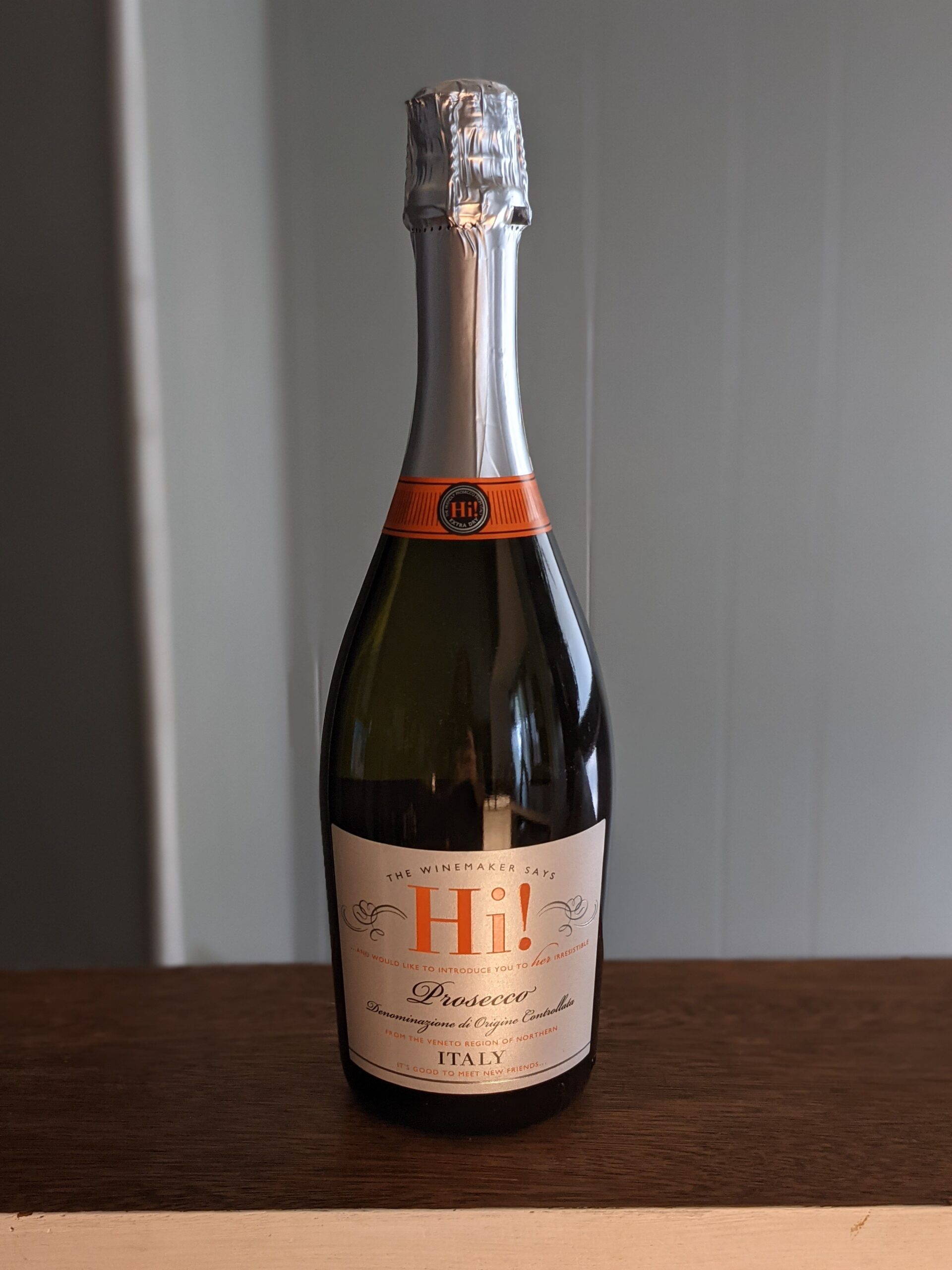 A sealed bottle of Hi! Prosecco on a wooden ledge, white panel wooden wall blurred in the background
