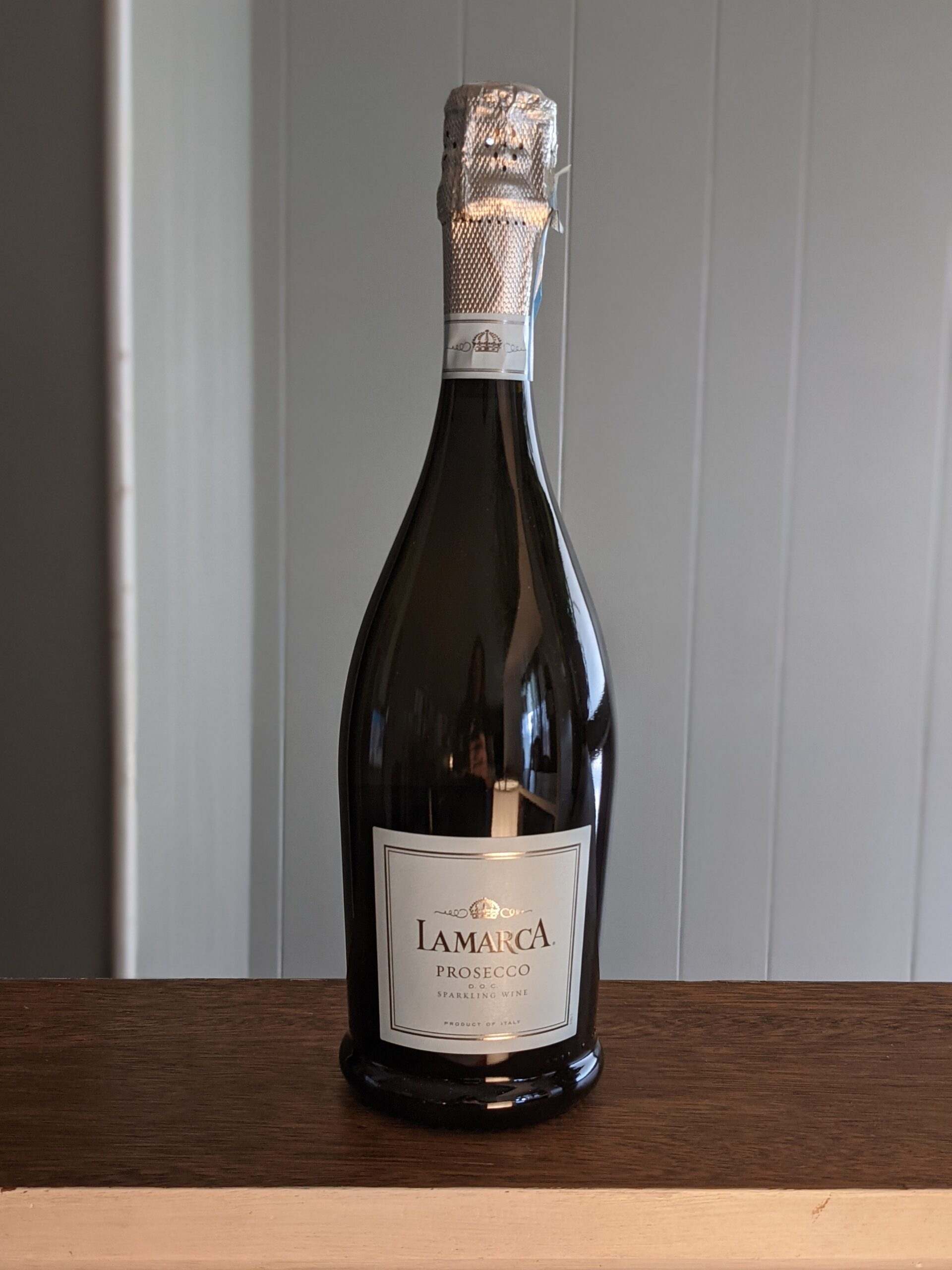 A sealed bottle of LaMarca Prosecco on a wooden ledge, white panel wooden wall blurred in the background