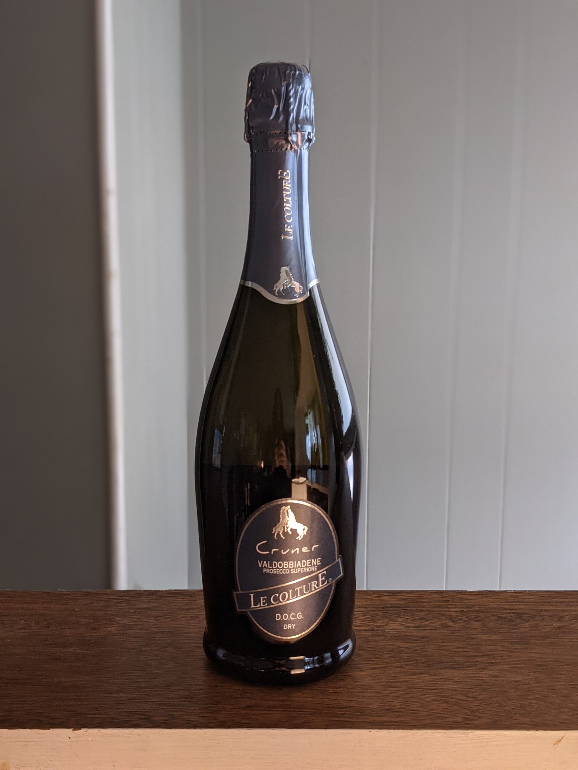 A sealed bottle of Le Colture Cruner Prosecco on a wooden ledge, white panel wooden wall blurred in the background