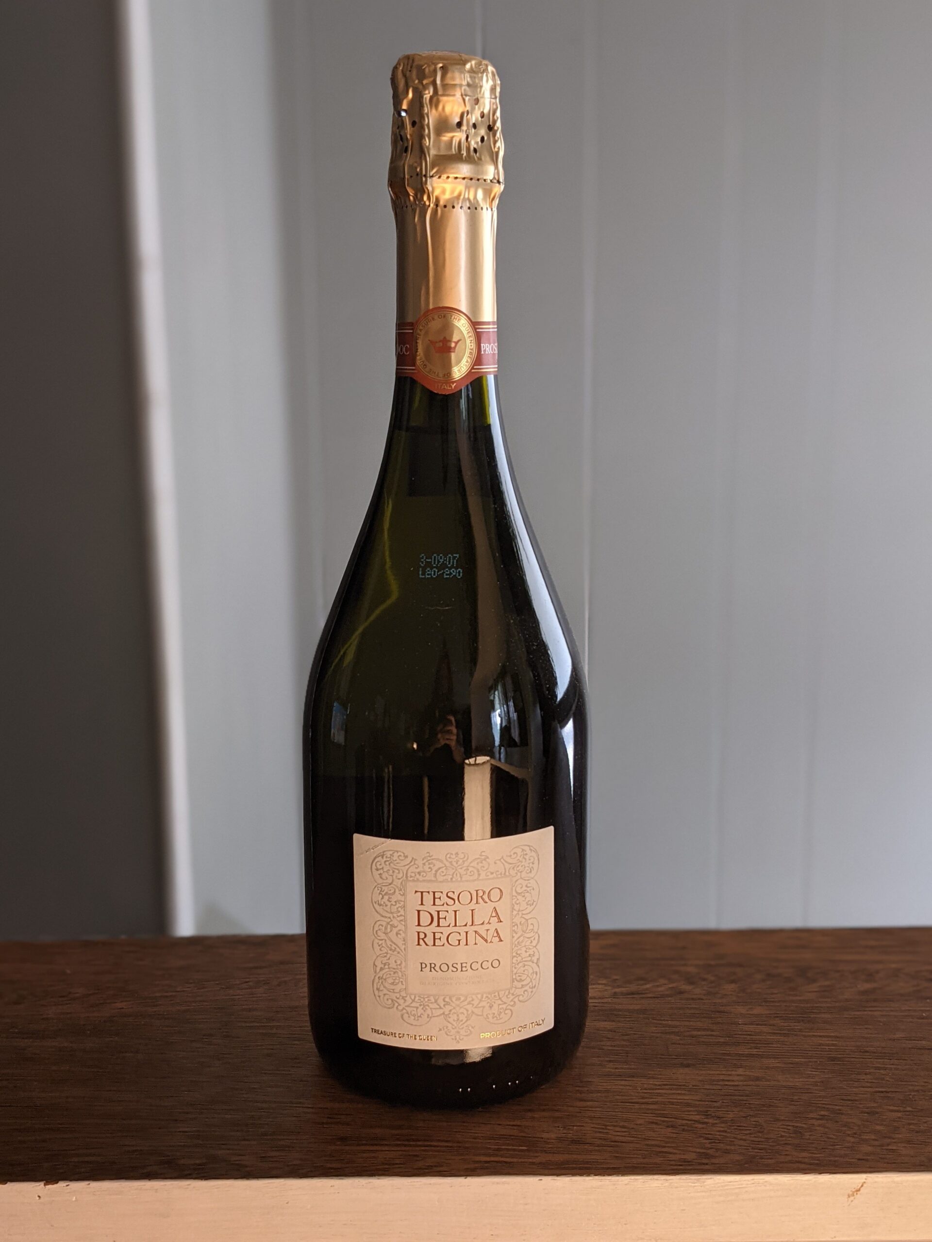 A sealed bottle of Tesoro Della Regina Prosecco on a wooden ledge, white panel wooden wall blurred in the background