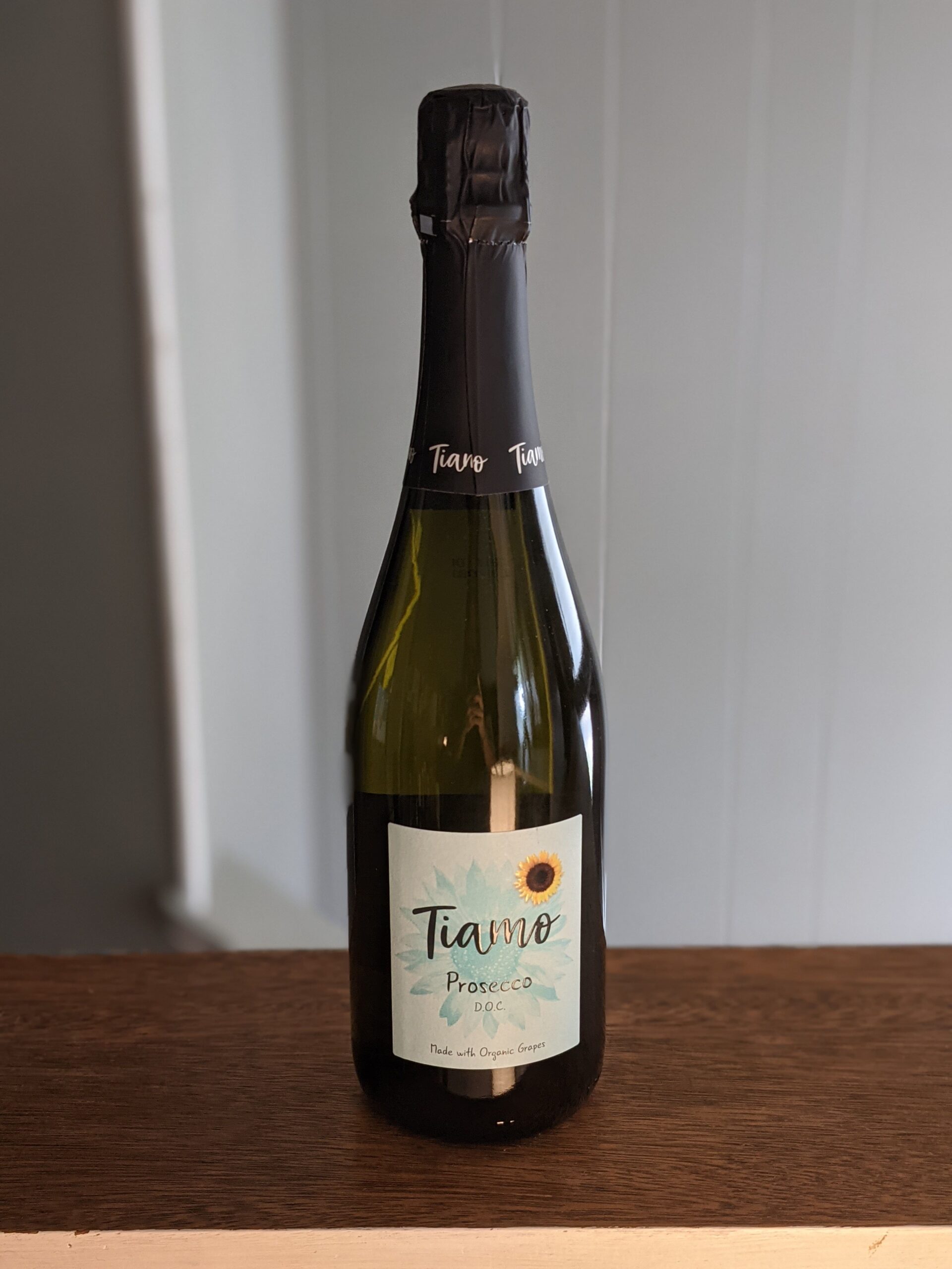 A sealed bottle of Tiamo Prosecco on a wooden ledge, white panel wooden wall blurred in the background