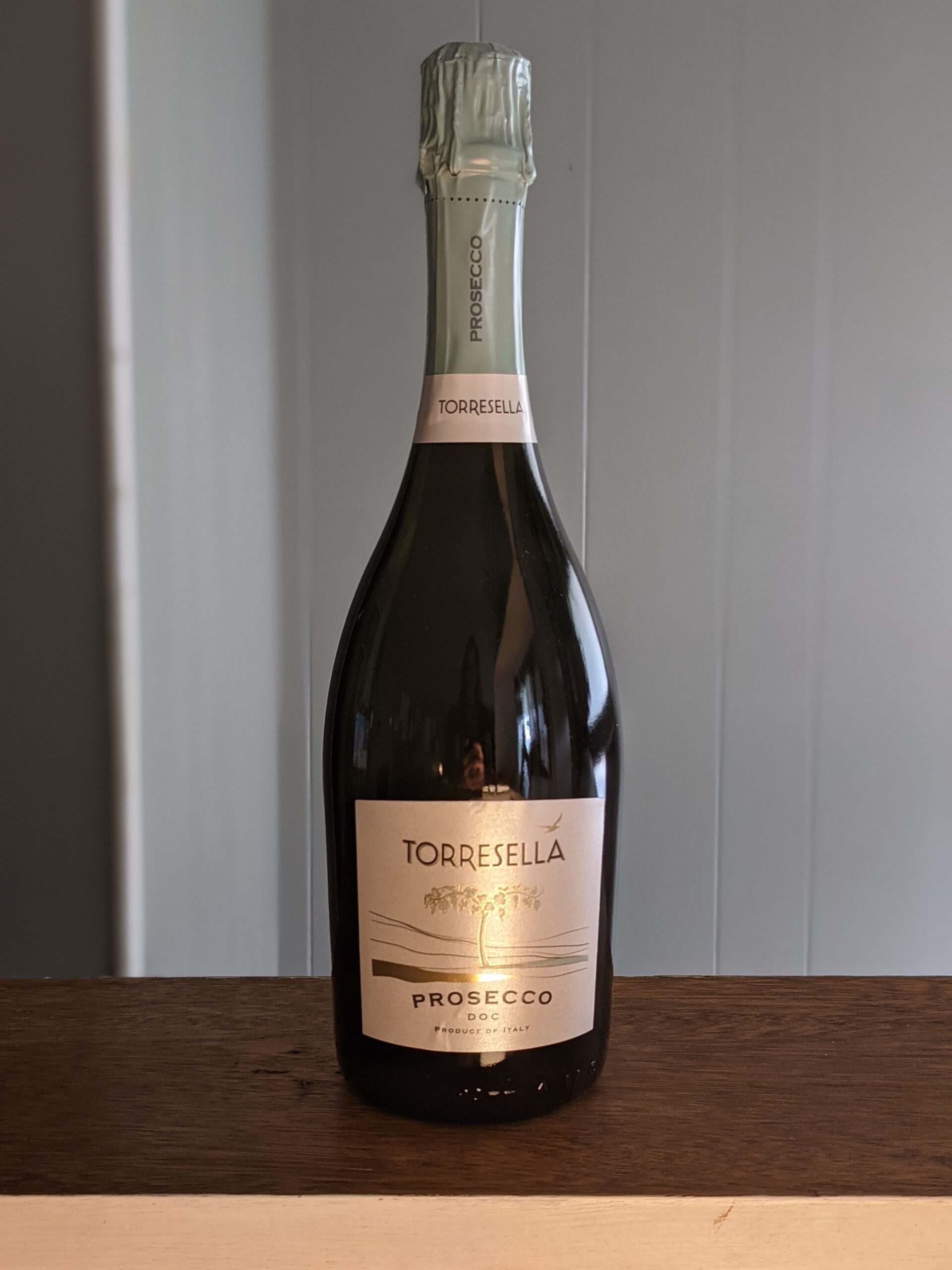 A sealed bottle of Torresella Prosecco on a wooden ledge, white panel wooden wall blurred in the background