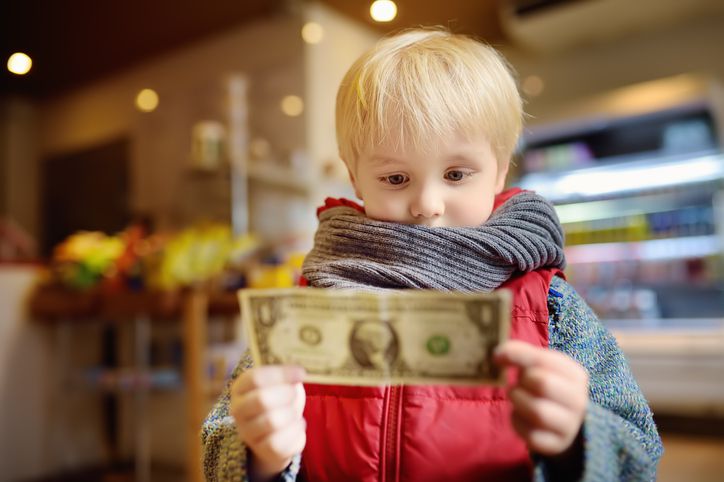 A young child wearing a scarf and red vest looks intently at a one-dollar bill while standing indoors, with a blurred background of a store or café.