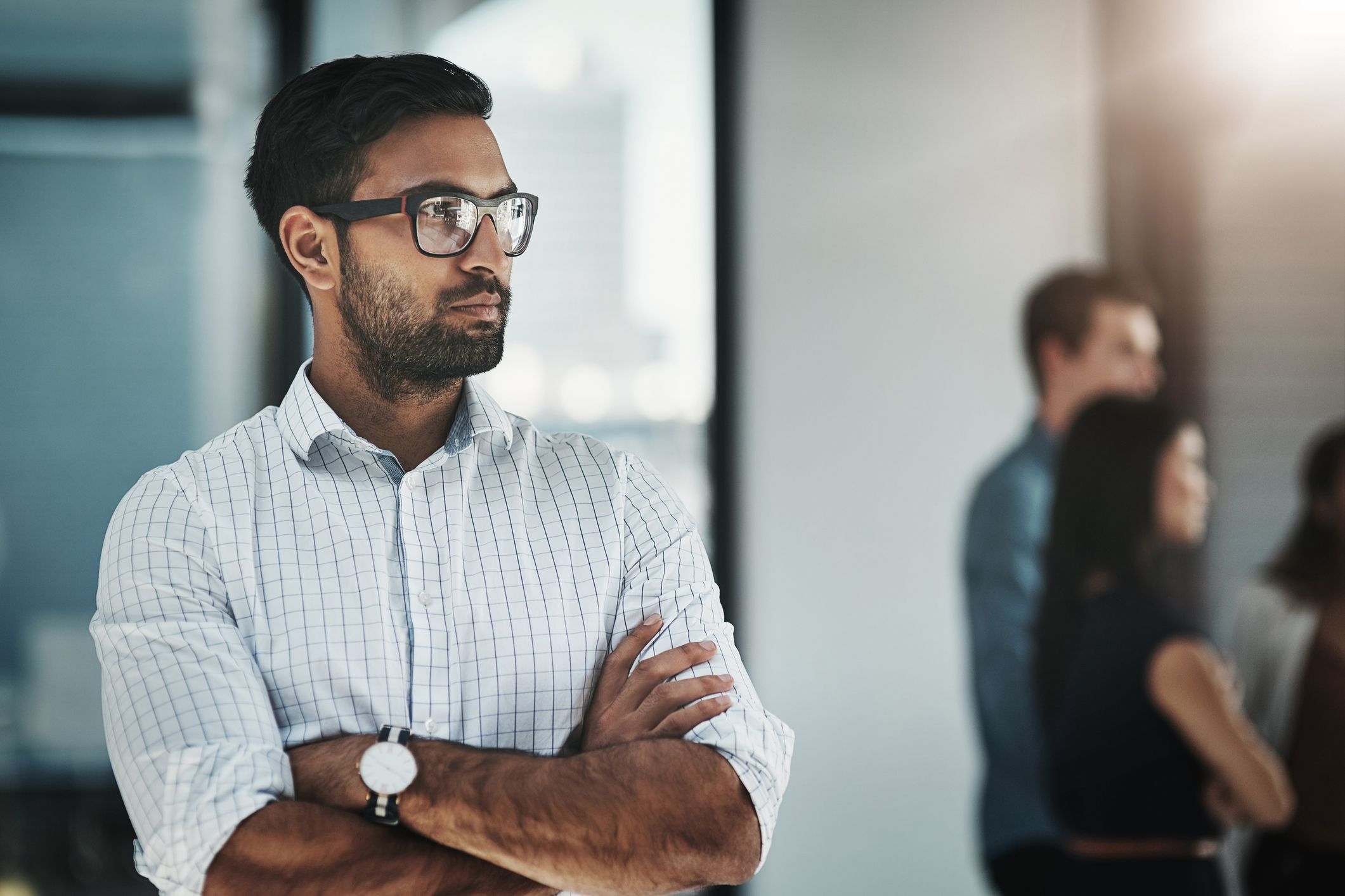 Shot of a young businessman posing with his arms folded in a modern office with his colleagues in the background