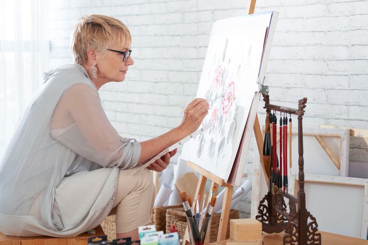 An older woman with short blonde hair and glasses sits on a stool, intently painting on a canvas at an easel in a bright art studio, surrounded by art supplies and tools.