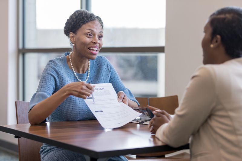 Two women sit across from each other at a table. One woman, smiling and wearing a blue dress, holds and gestures to a job application form while talking to the other woman, who is listening attentively.