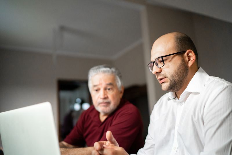 Two men, one older with gray hair and a beard, and one younger with glasses and a bald head, sit together indoors, looking at a laptop screen with serious expressions.