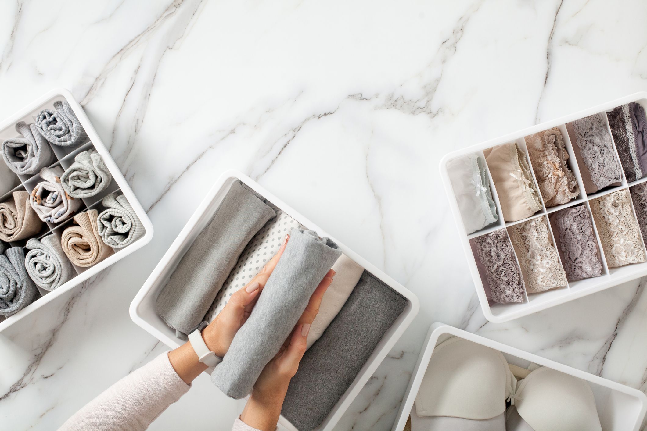 A person organizes folded clothes, socks, and underwear in white storage bins on a marble surface, arranging items neatly by color and type for a tidy drawer.