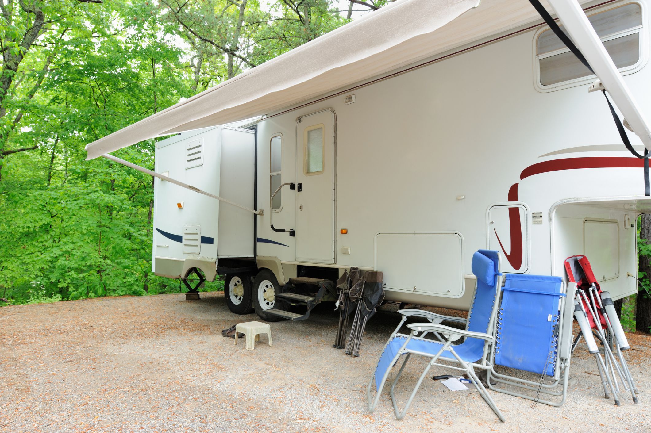 Recreational vehicle fifth wheel travel trailer in wooded campsite with awning and folding chairs.