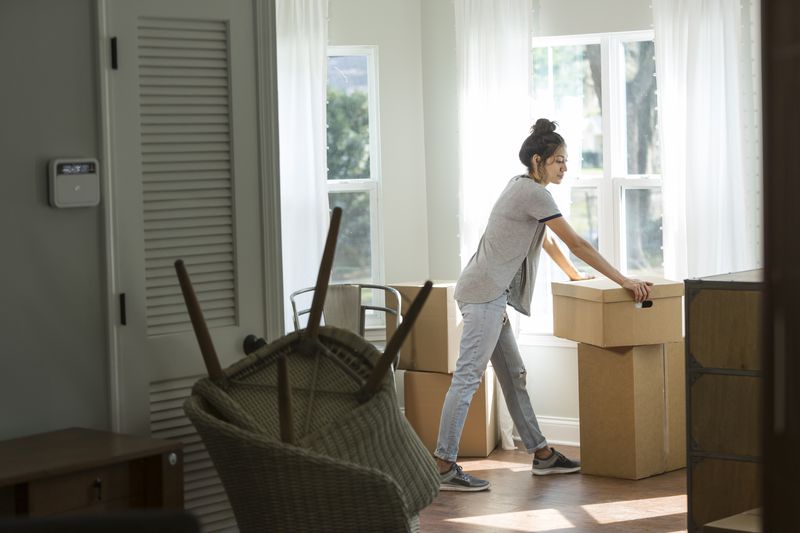 A woman stands in a sunlit room pushing a cardboard box, surrounded by other moving boxes and baskets, with large windows in the background.