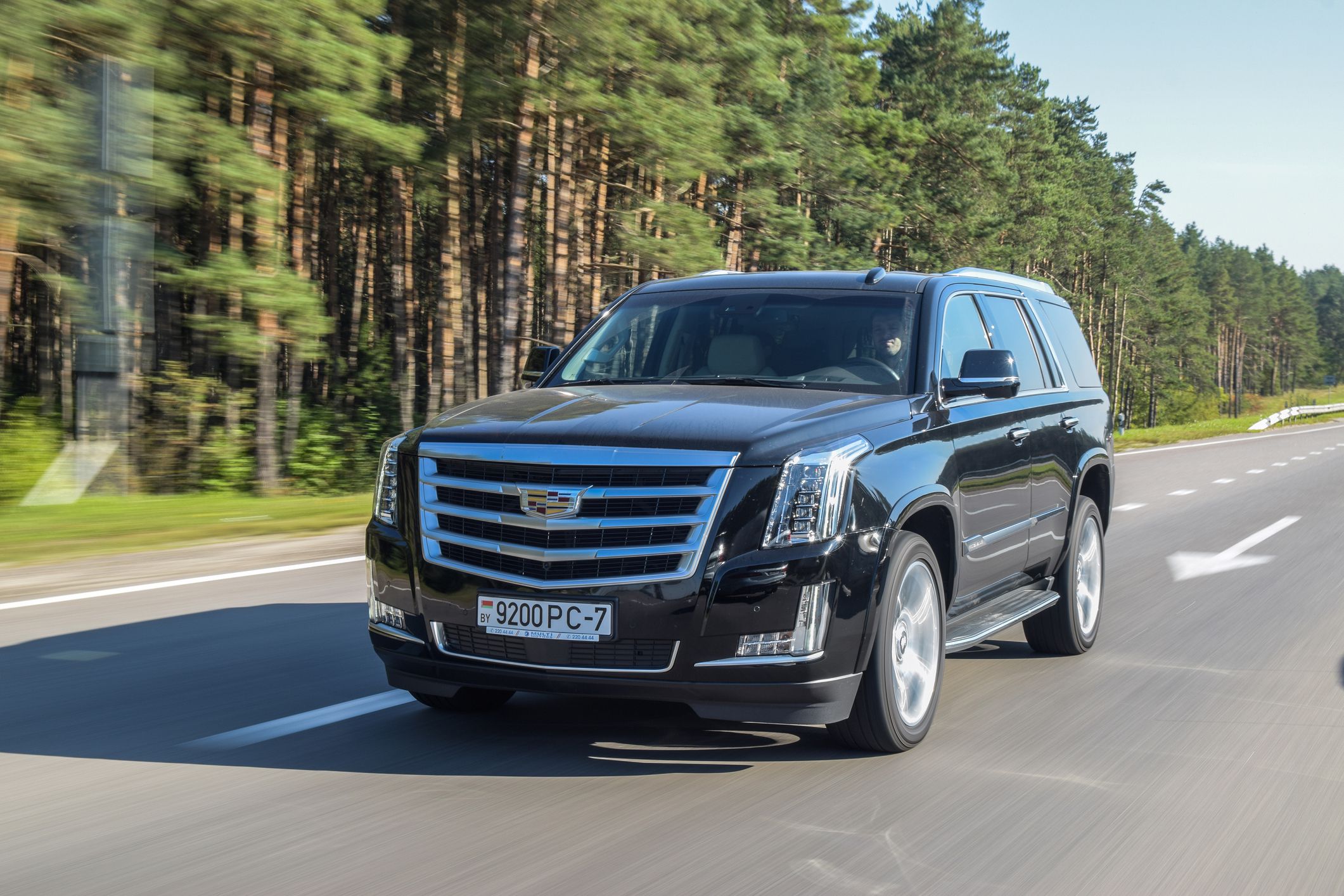 A black Cadillac Escalade SUV driving on a multi-lane highway surrounded by trees and greenery on a clear day.
