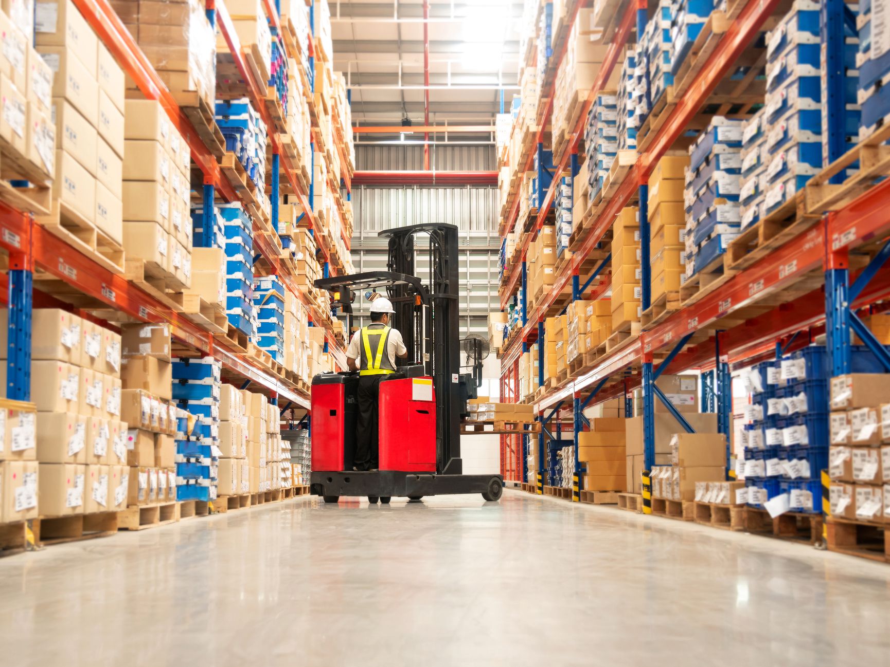 Worker in forklift-truck loading packed goods in huge distribution warehouse with high shelves.