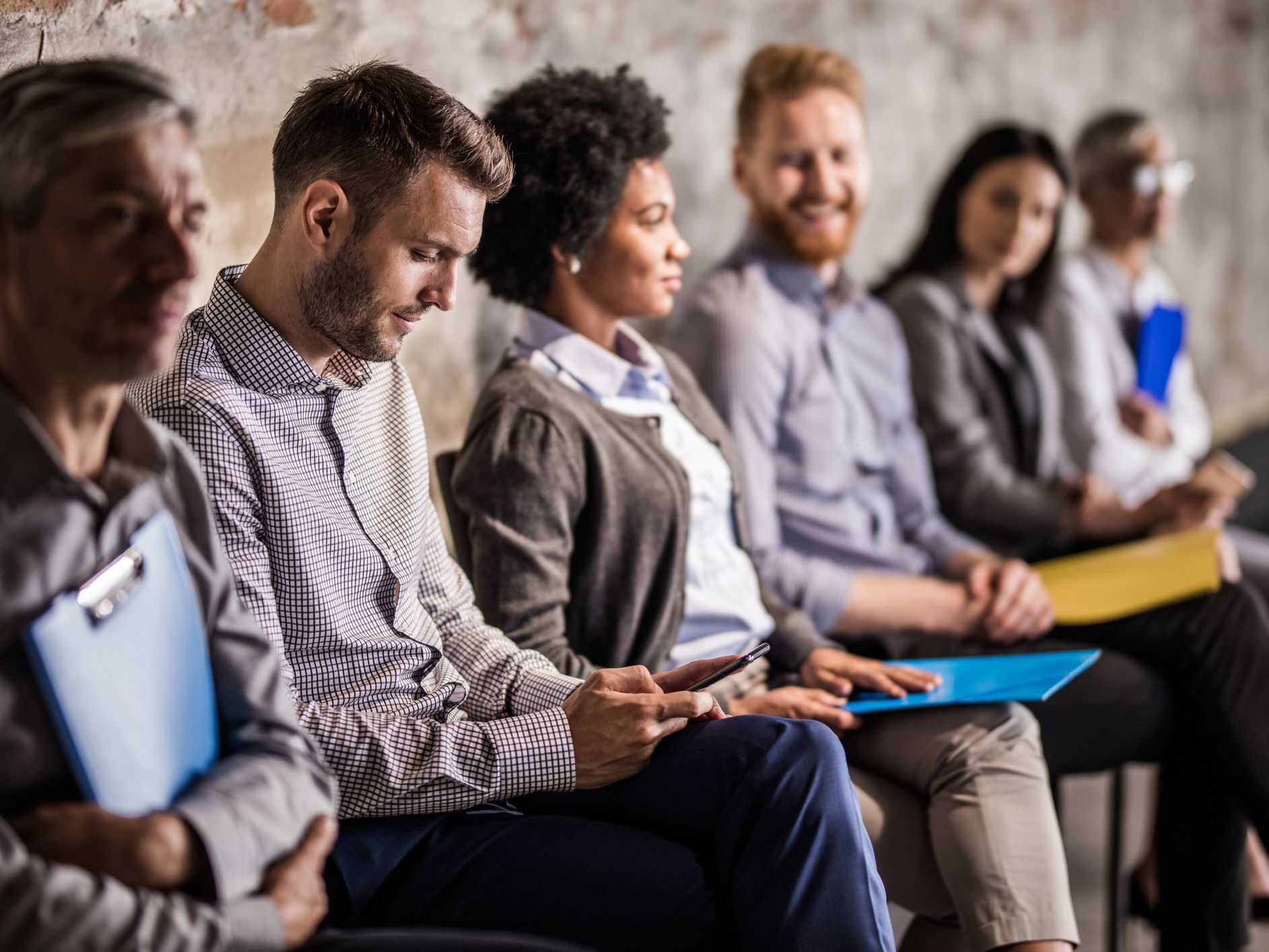 Large group of business people sitting in waiting room before their job interview. Focus is on man text messaging on cell phone.