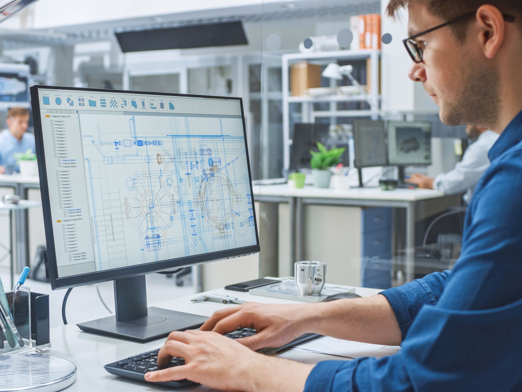 Over the Shoulder Shot of Engineer Working with CAD Software on Desktop Computer, Screen Shows Technical Drafts and Drawings. In the Background Engineering Facility Specialising on Industrial Design