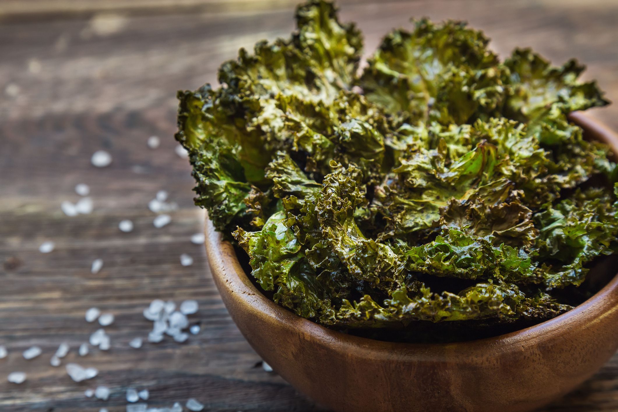 A wooden bowl filled with crispy baked kale chips sits on a wooden surface, with coarse salt scattered around.