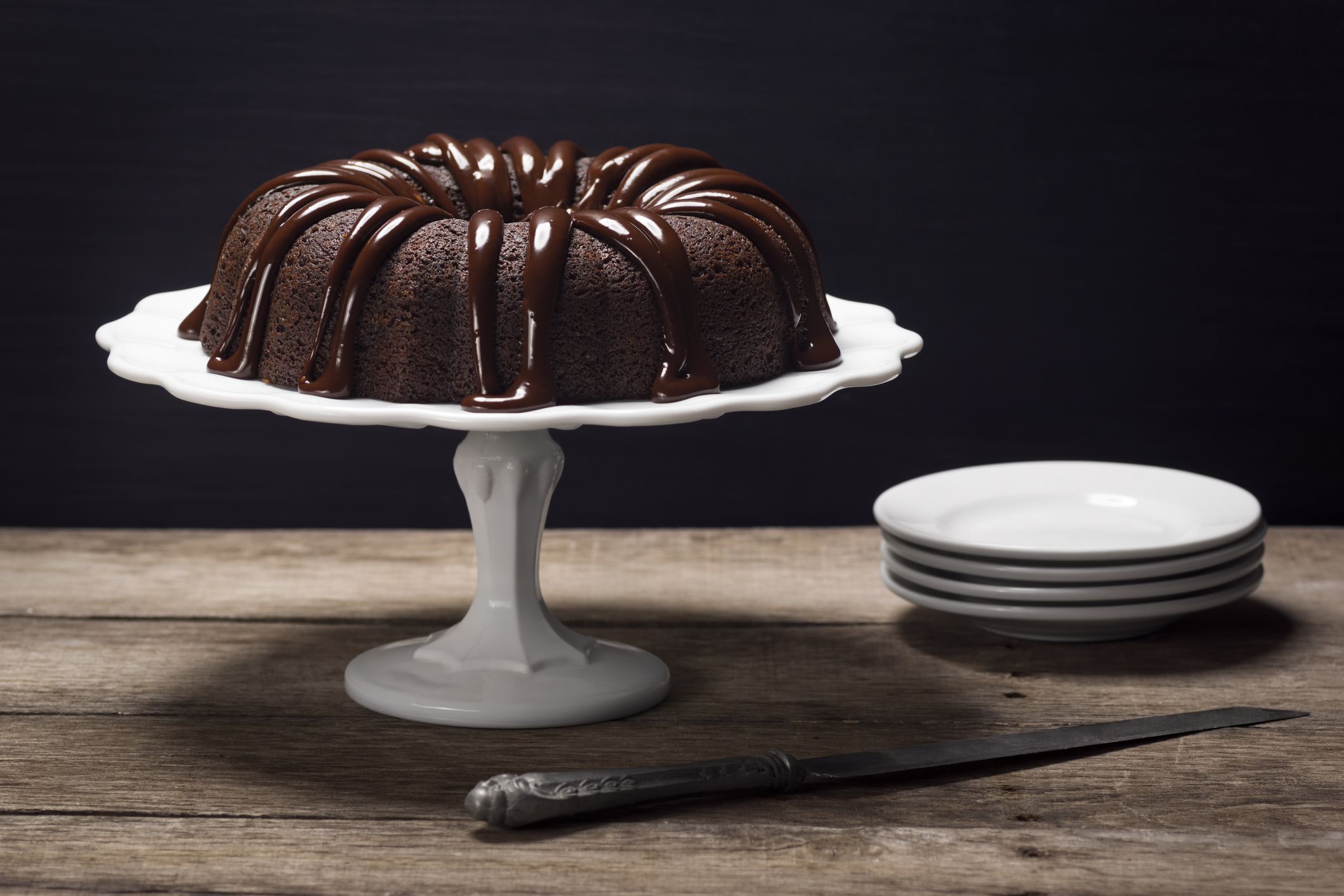 A chocolate bundt cake with ganache icing sits atop a cake stand on a wooden table.