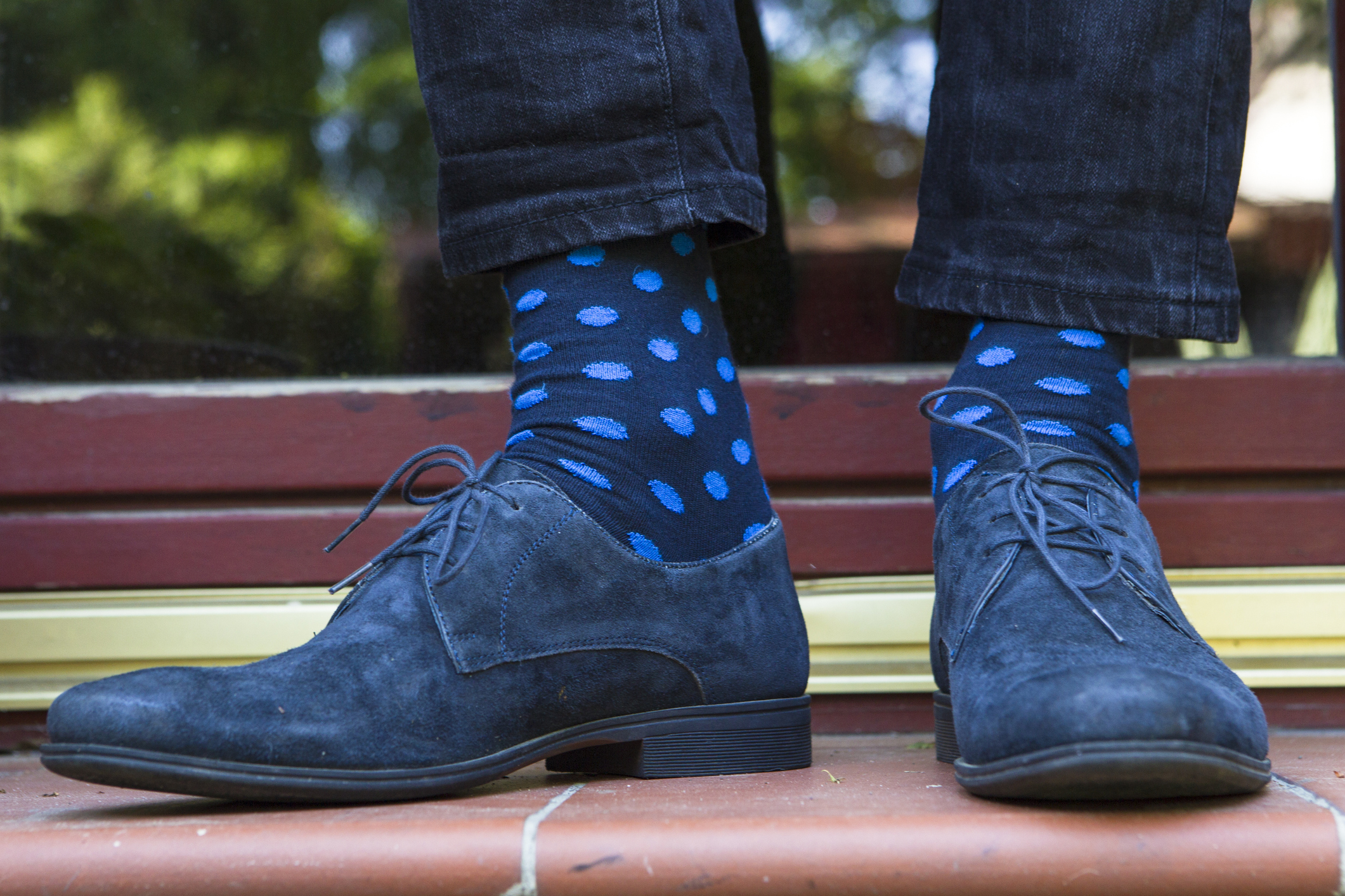 Closeup of a blue suede shoes with matching cobalt blue polka-dots on navy socks, man wearing with a bit of jeans, on dark red tiles with a blurred background of bushes and yard